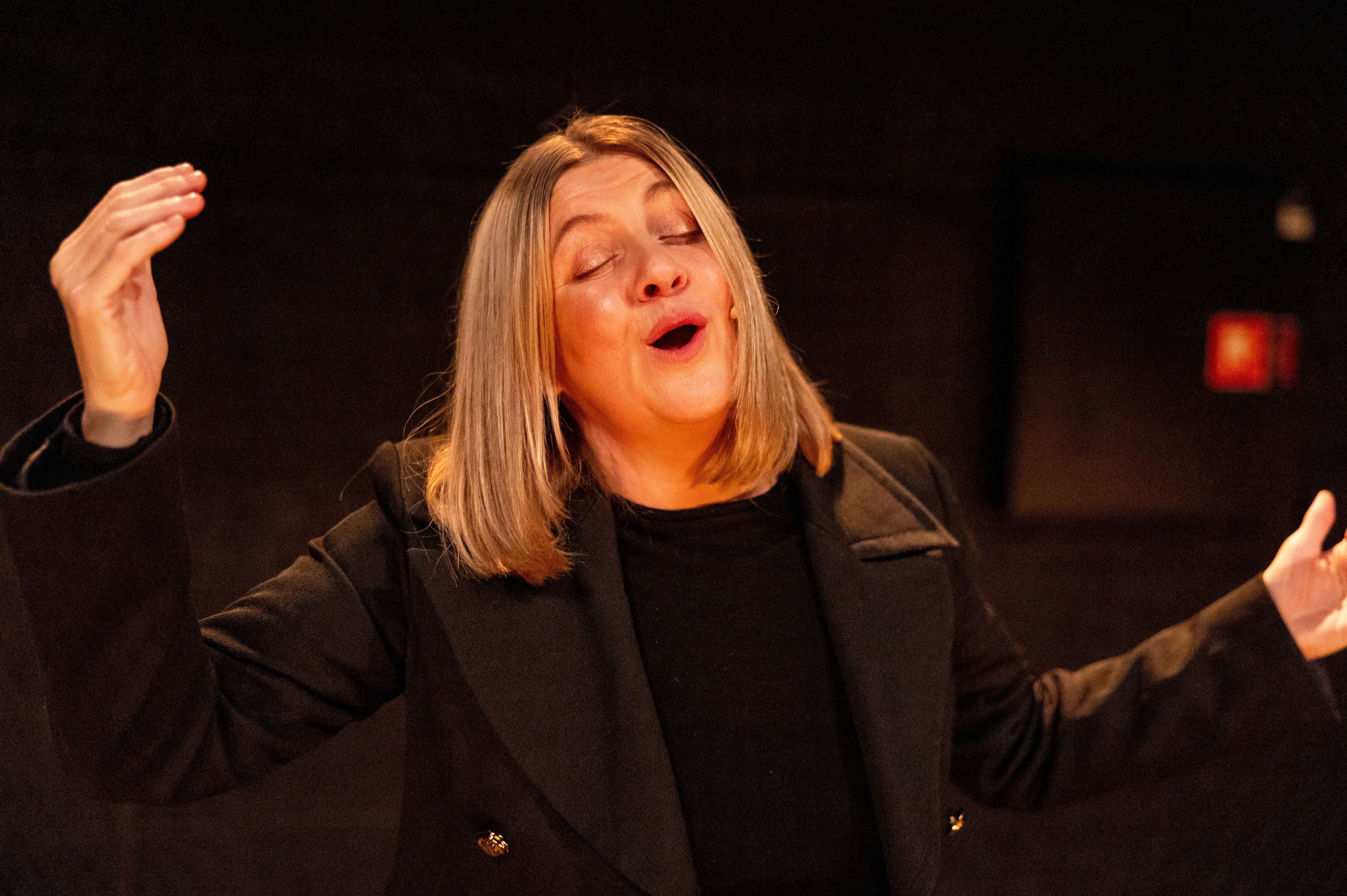 Kvinne med skulderlangt blondt hår i svart antrekk gestikulerer med lukkede øyne under scenelys.