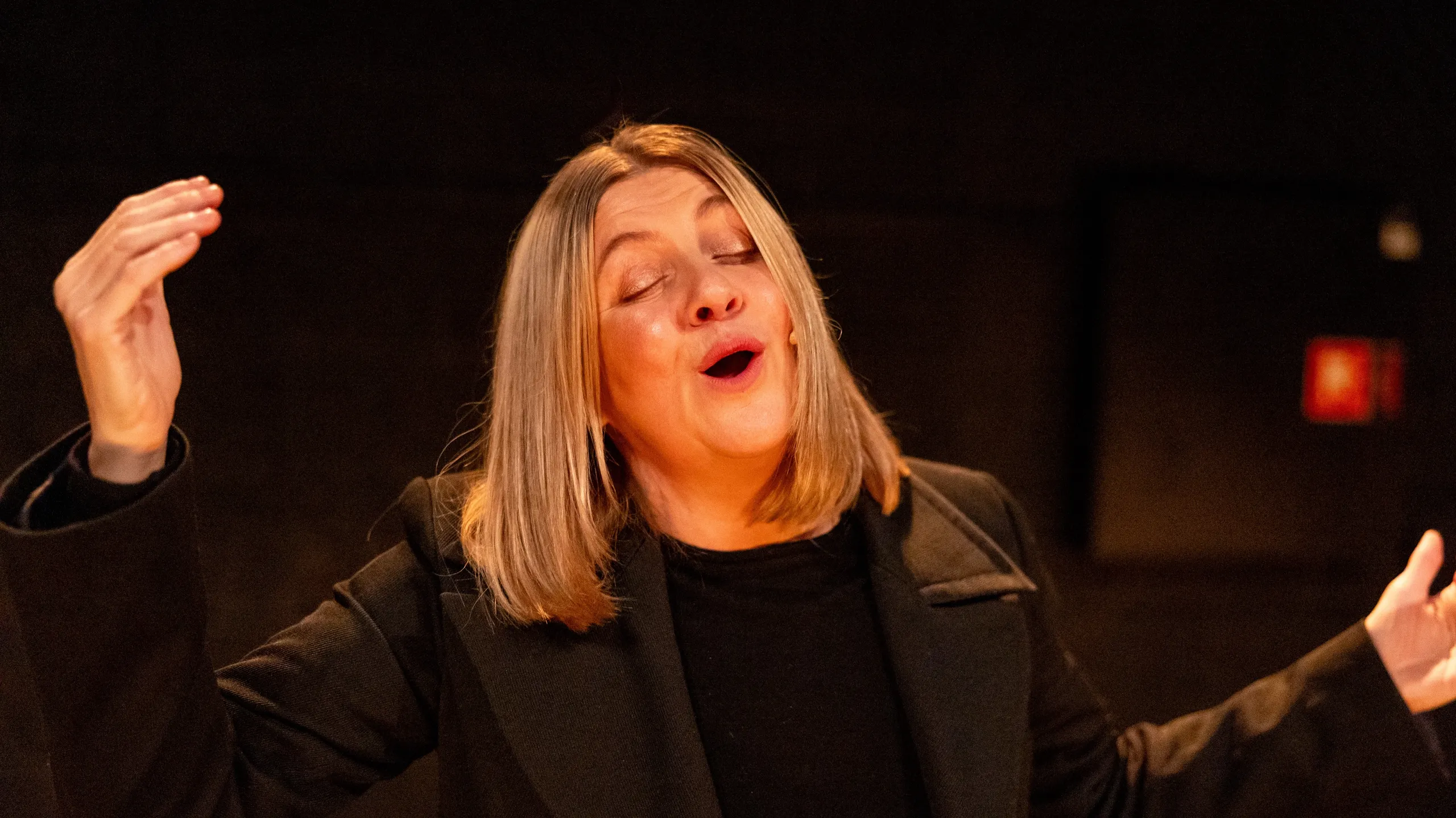 Kvinne med skulderlangt blondt hår i svart antrekk gestikulerer med lukkede øyne under scenelys.