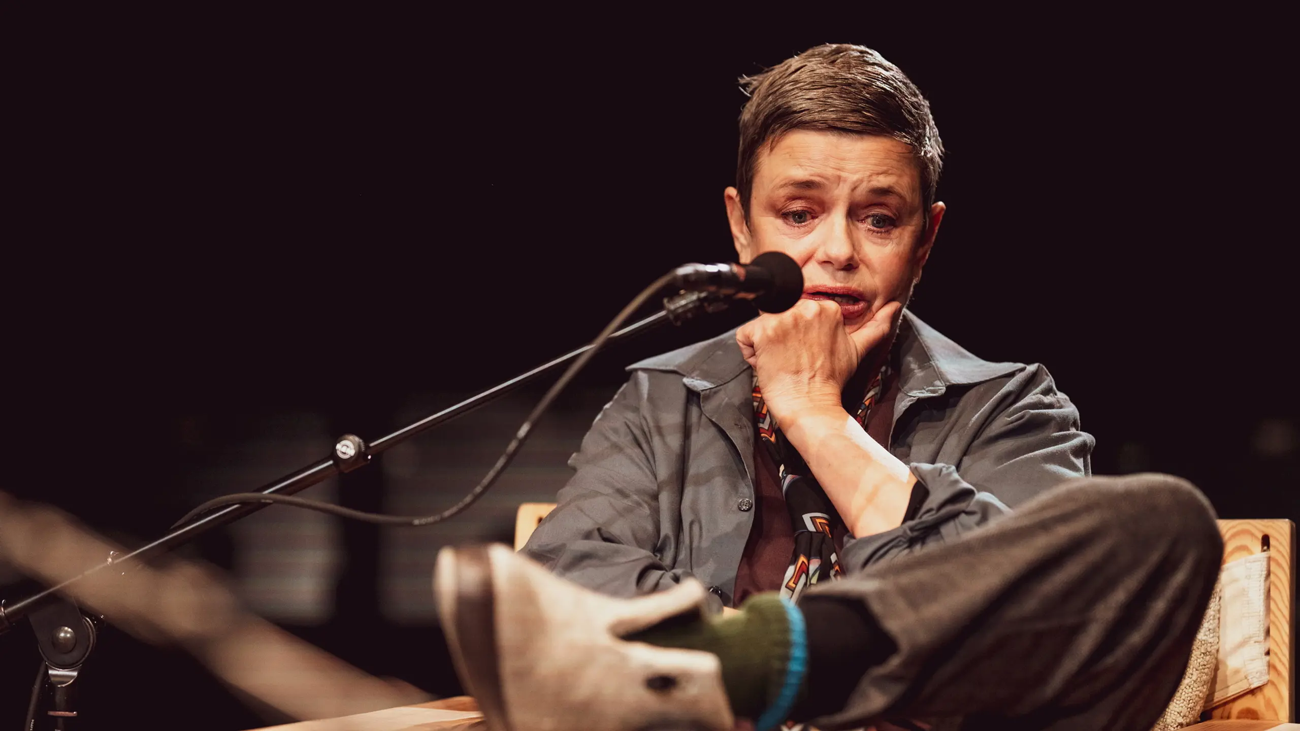 Person med kort hår sitter tankefullt med hånden mot haken og foten hvilende på stolarmen.