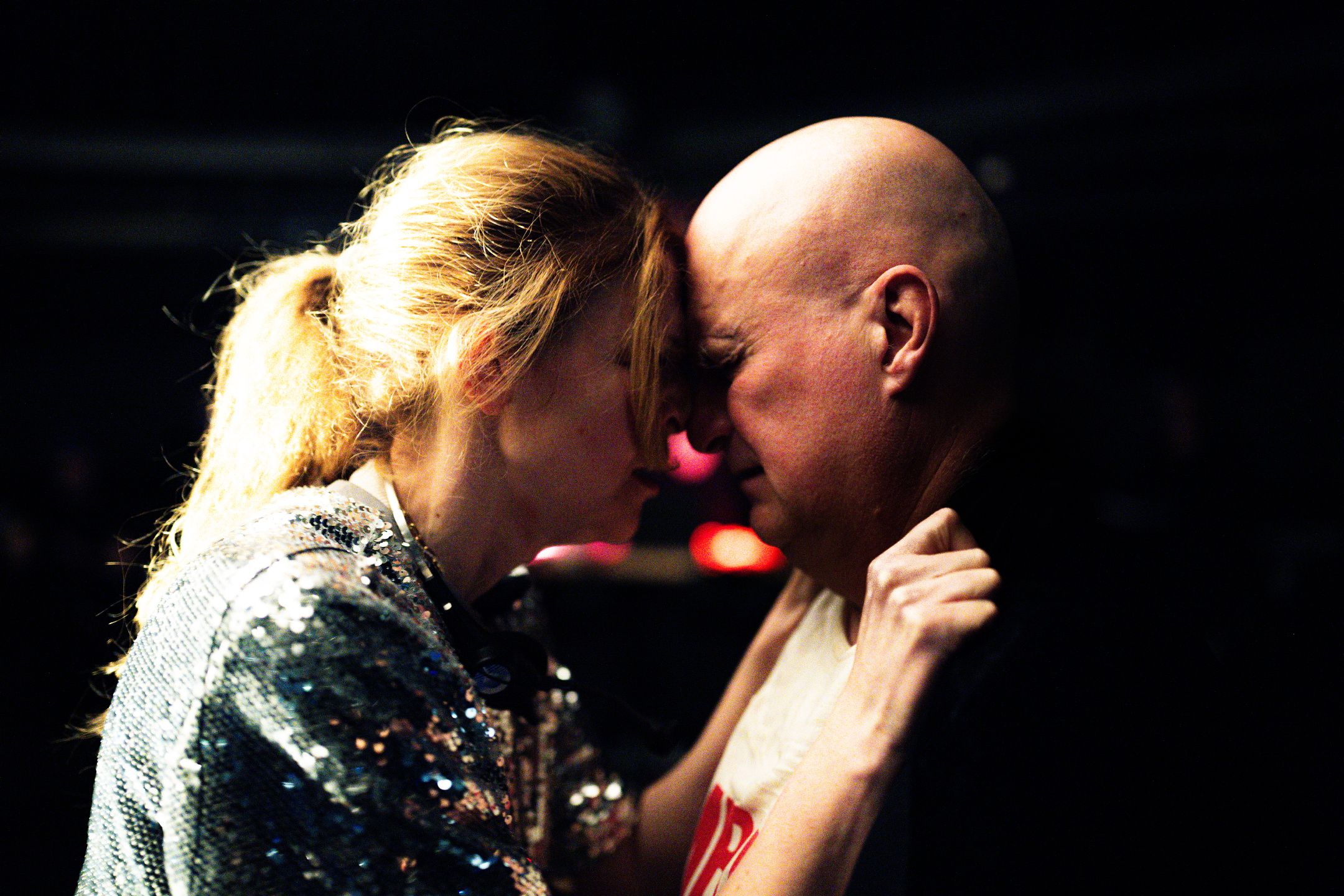 Kvinne og mann står panne mot panne i mørkt rom, kvinnen holder tak i jakkekragen hans