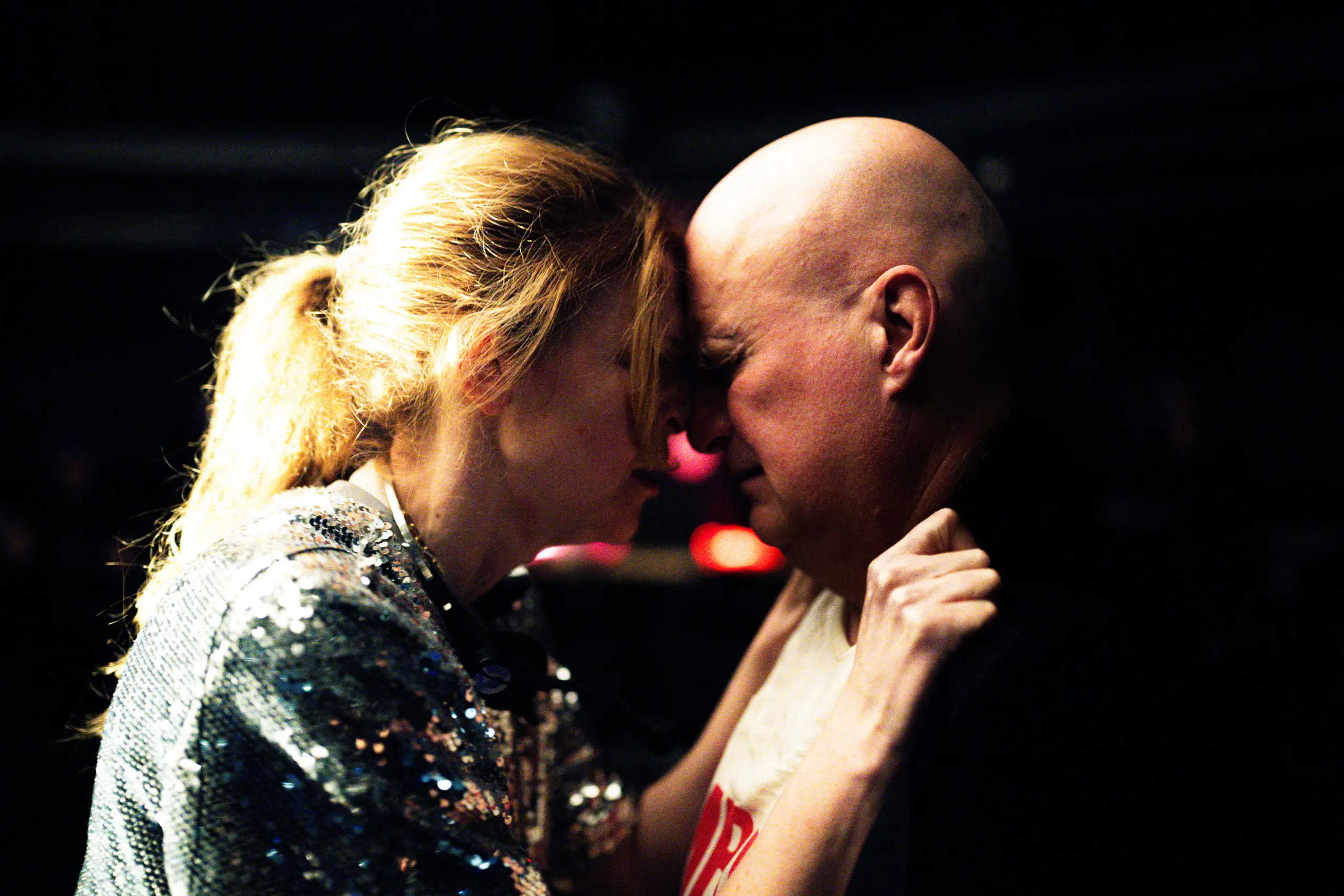 Kvinne og mann står panne mot panne i mørkt rom, kvinnen holder tak i jakkekragen hans