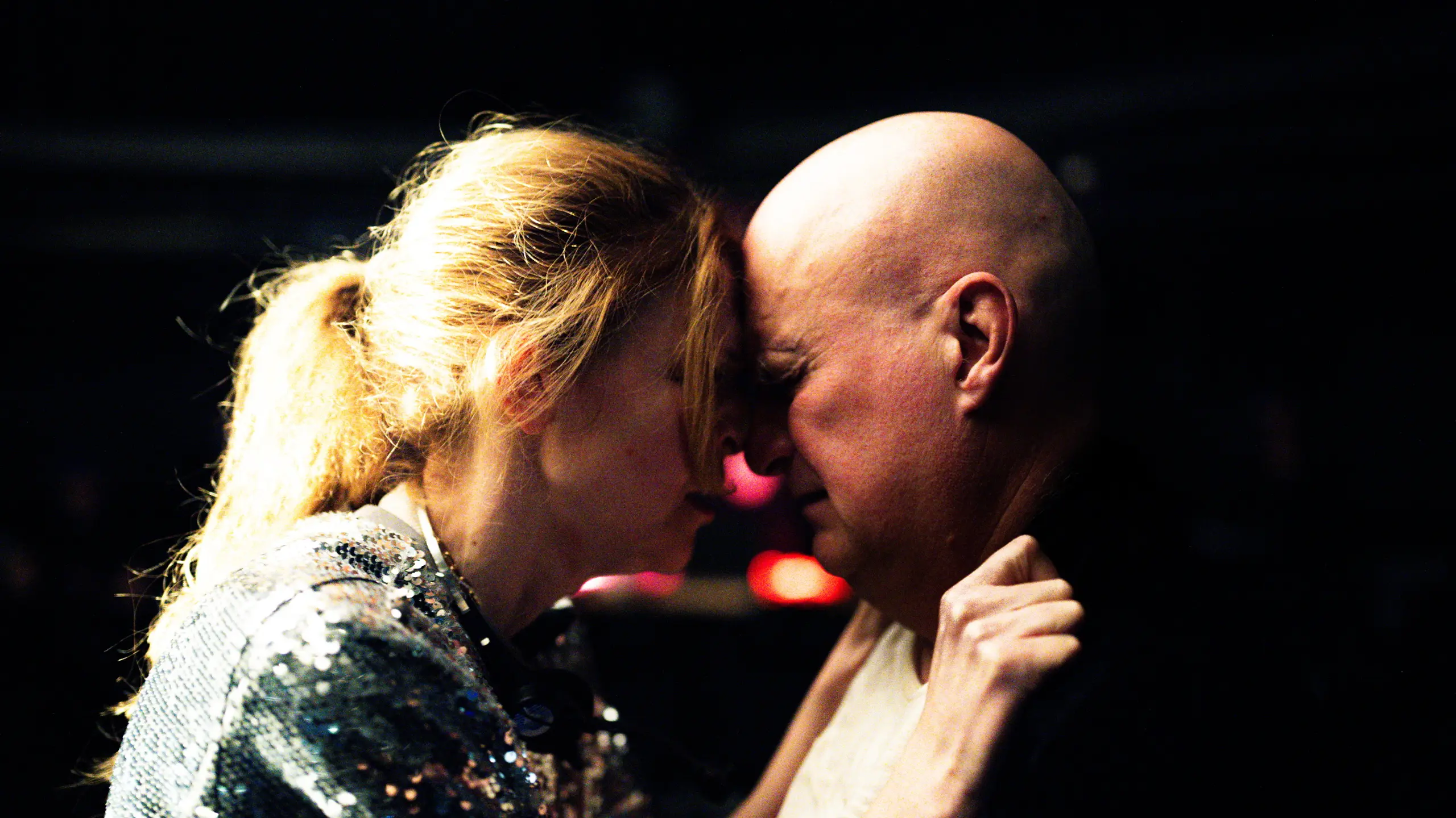 Kvinne og mann står panne mot panne i mørkt rom, kvinnen holder tak i jakkekragen hans