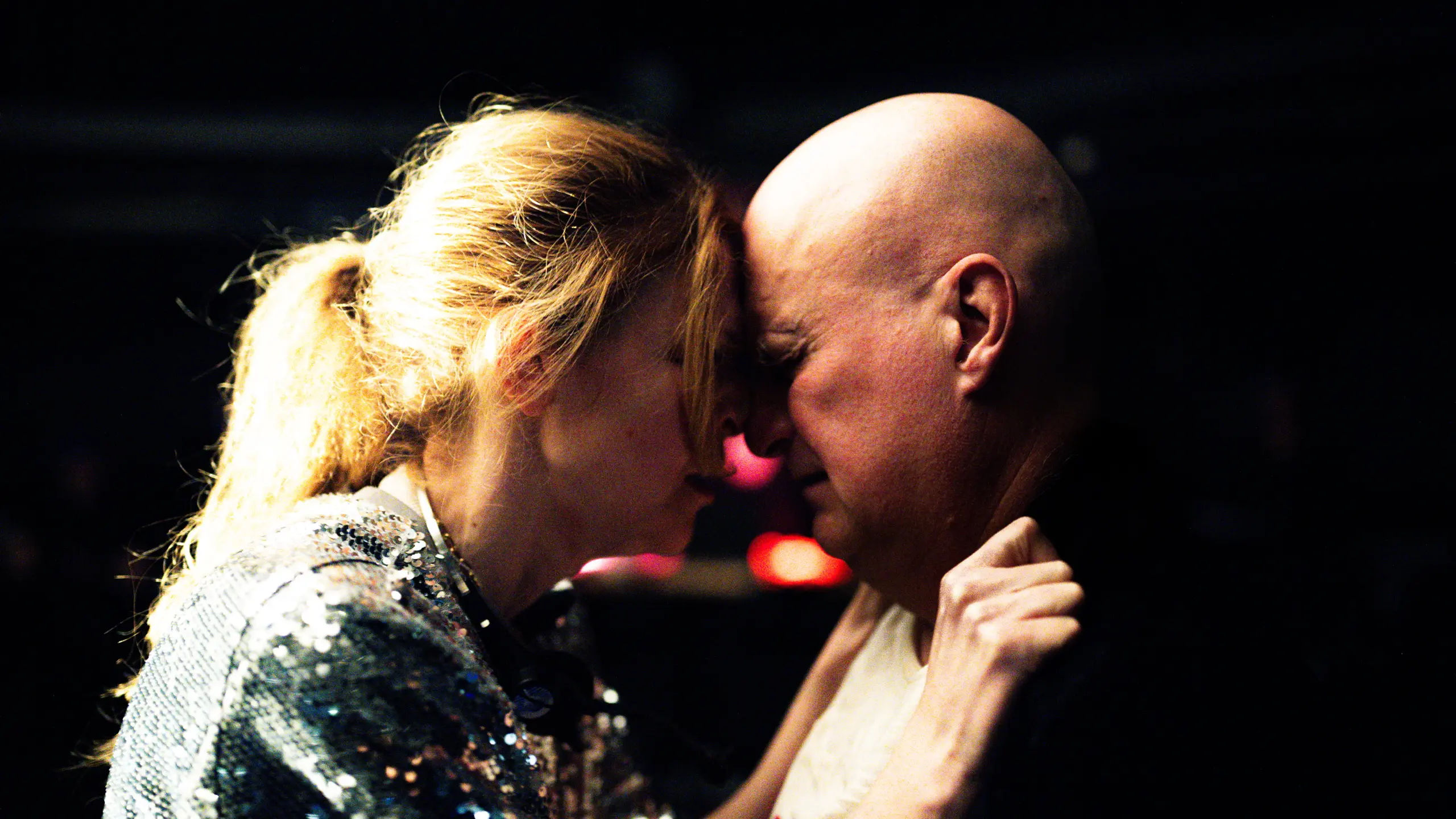 Kvinne og mann står panne mot panne i mørkt rom, kvinnen holder tak i jakkekragen hans