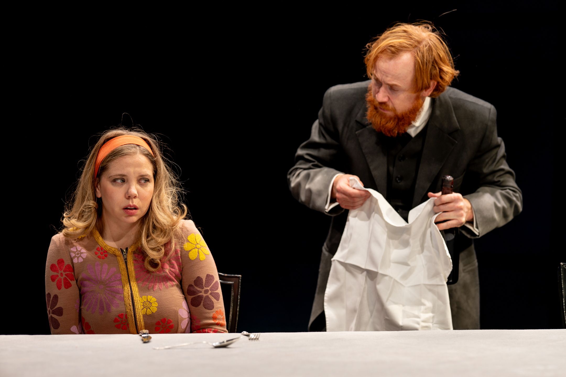 Kvinne i blomstret cardigan sitter ved bord og ser redd ut mens en mann i sort dress holder en hvit duk bak henne.