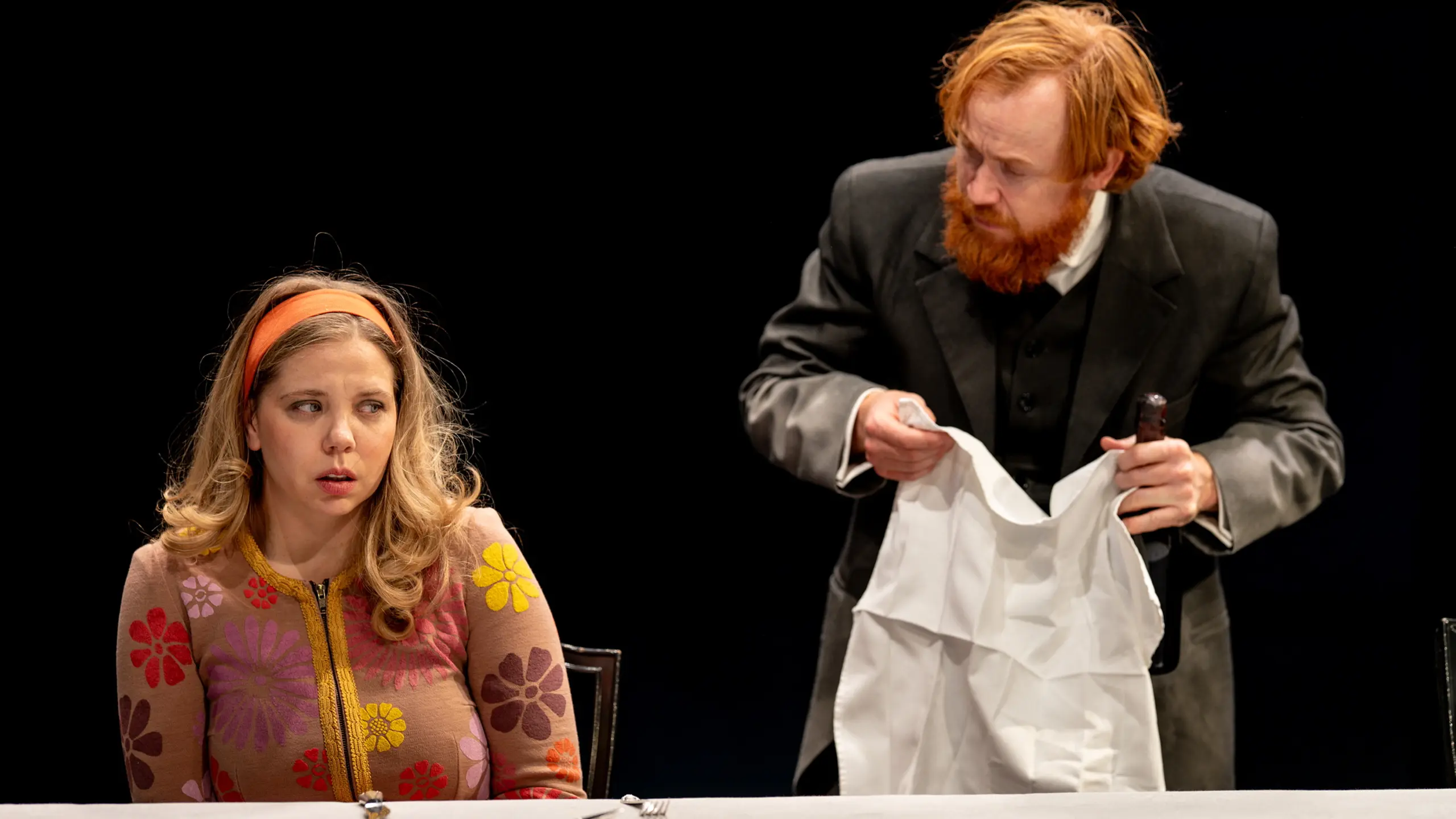 Kvinne i blomstret cardigan sitter ved bord og ser redd ut mens en mann i sort dress holder en hvit duk bak henne.