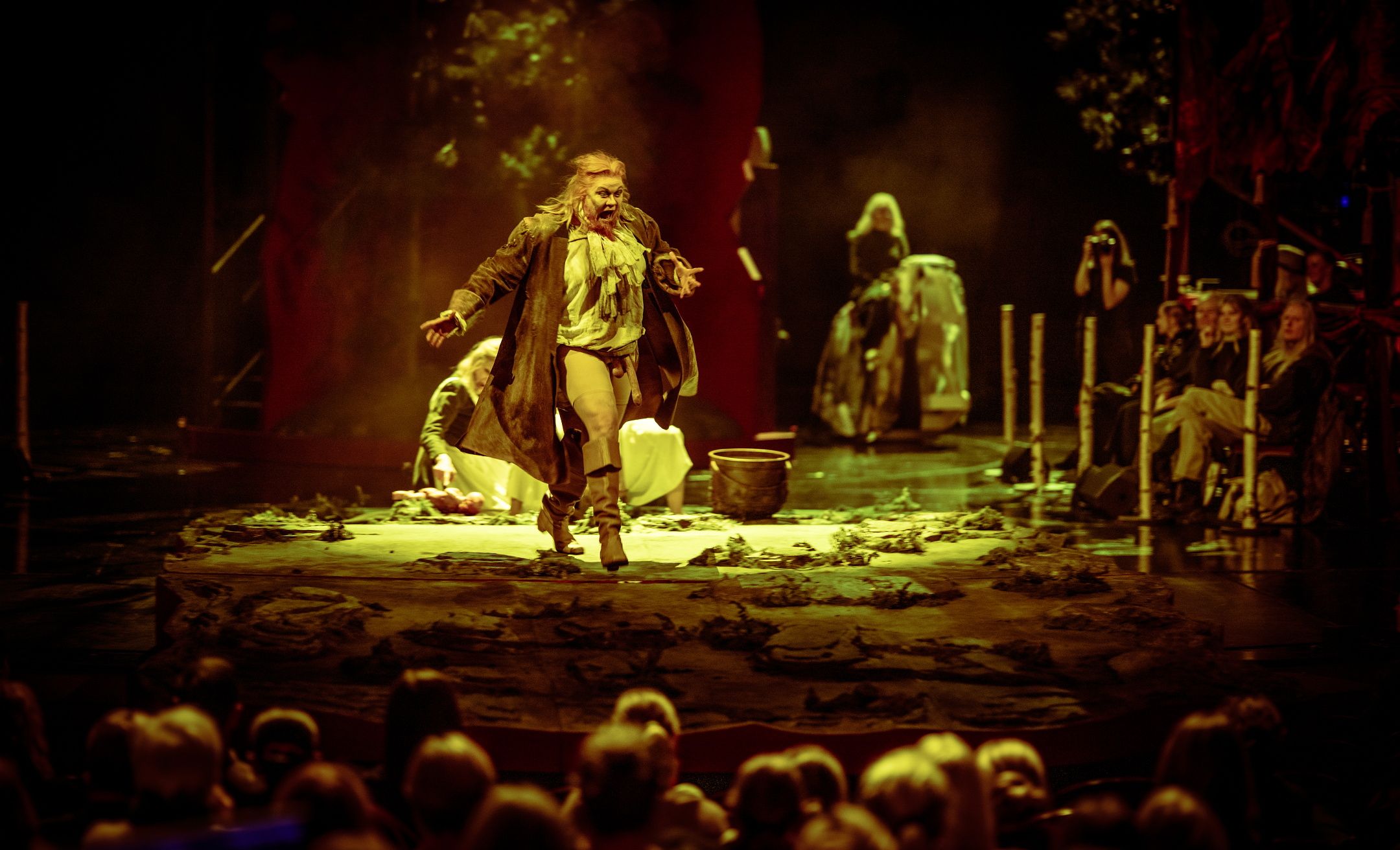 En skuespiller med langt hår og teatralsk kostyme roper mens hun løper mot publikum på en mørklagt scene med jordgulv.