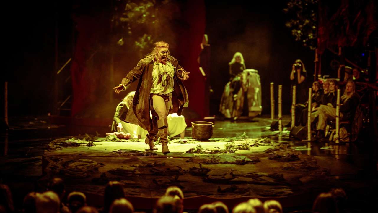 En skuespiller med langt hår og teatralsk kostyme roper mens hun løper mot publikum på en mørklagt scene med jordgulv.