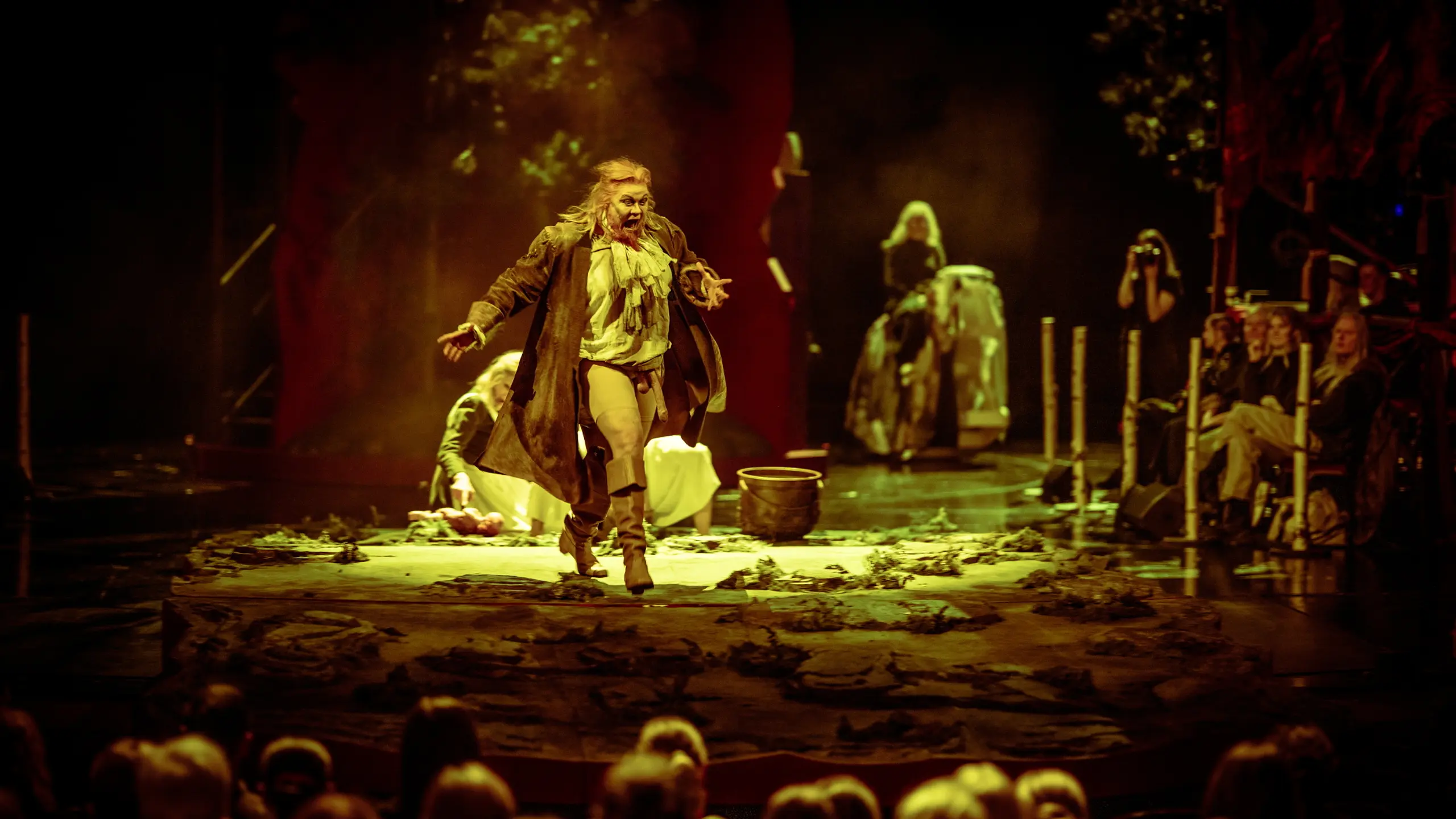 En skuespiller med langt hår og teatralsk kostyme roper mens hun løper mot publikum på en mørklagt scene med jordgulv.