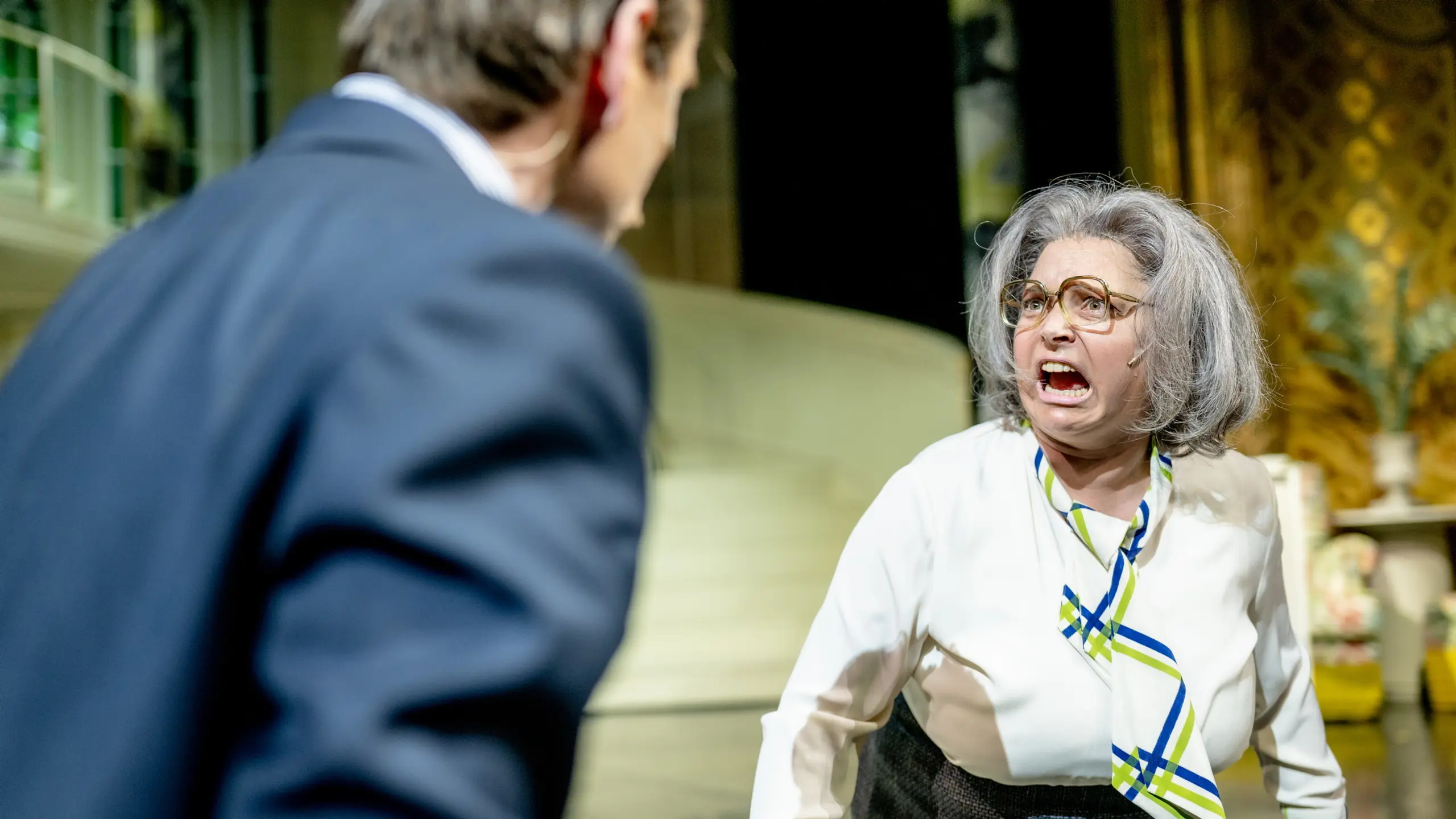 Kvinne med grå parykk og briller roper mot mann i dress på teaterscene