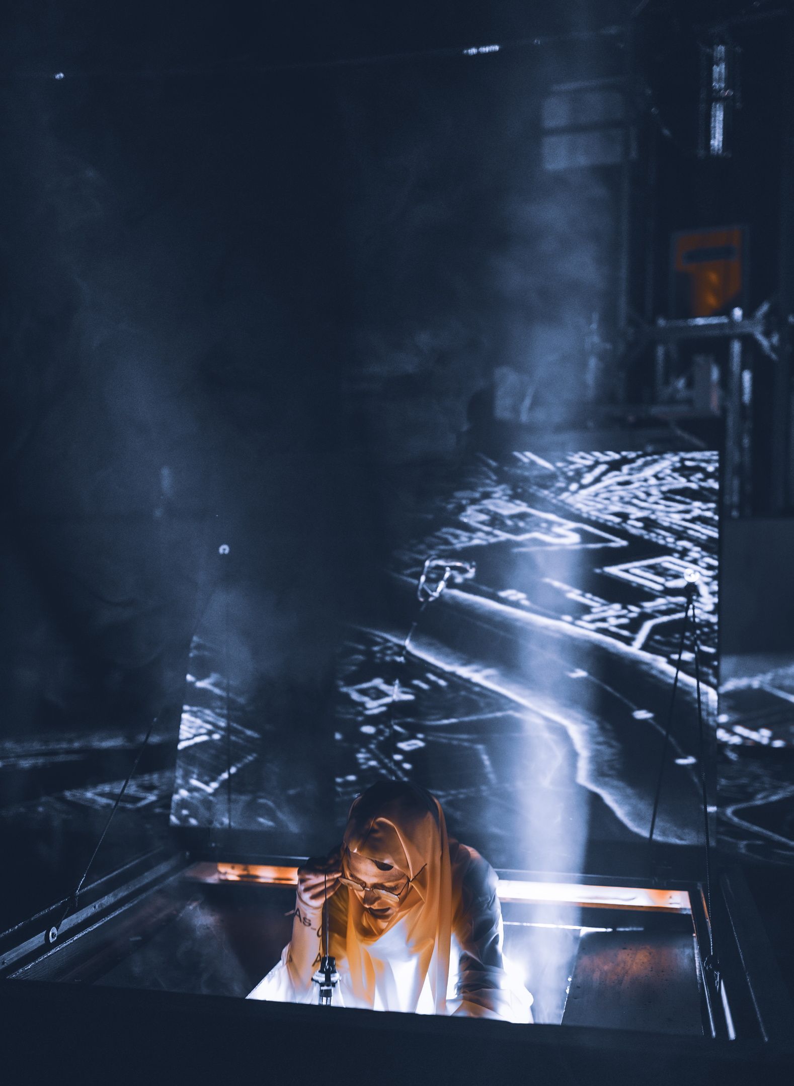 Et sceneelement åpner seg som en luke i gulvet, og en person med kappe og briller stiger opp i dramatisk lys. Røyk, mørke og projeksjoner