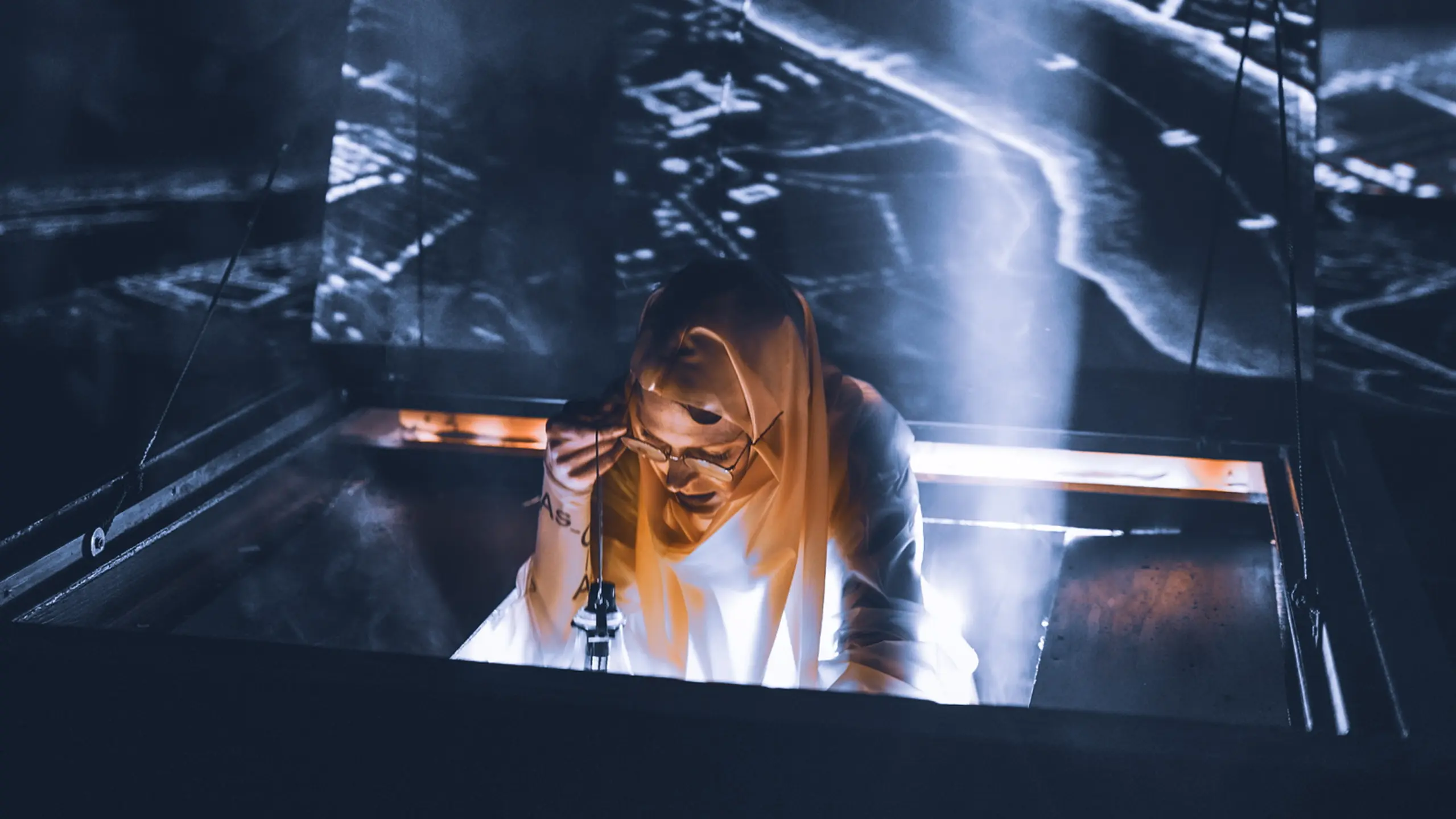 Et sceneelement åpner seg som en luke i gulvet, og en person med kappe og briller stiger opp i dramatisk lys. Røyk, mørke og projeksjoner