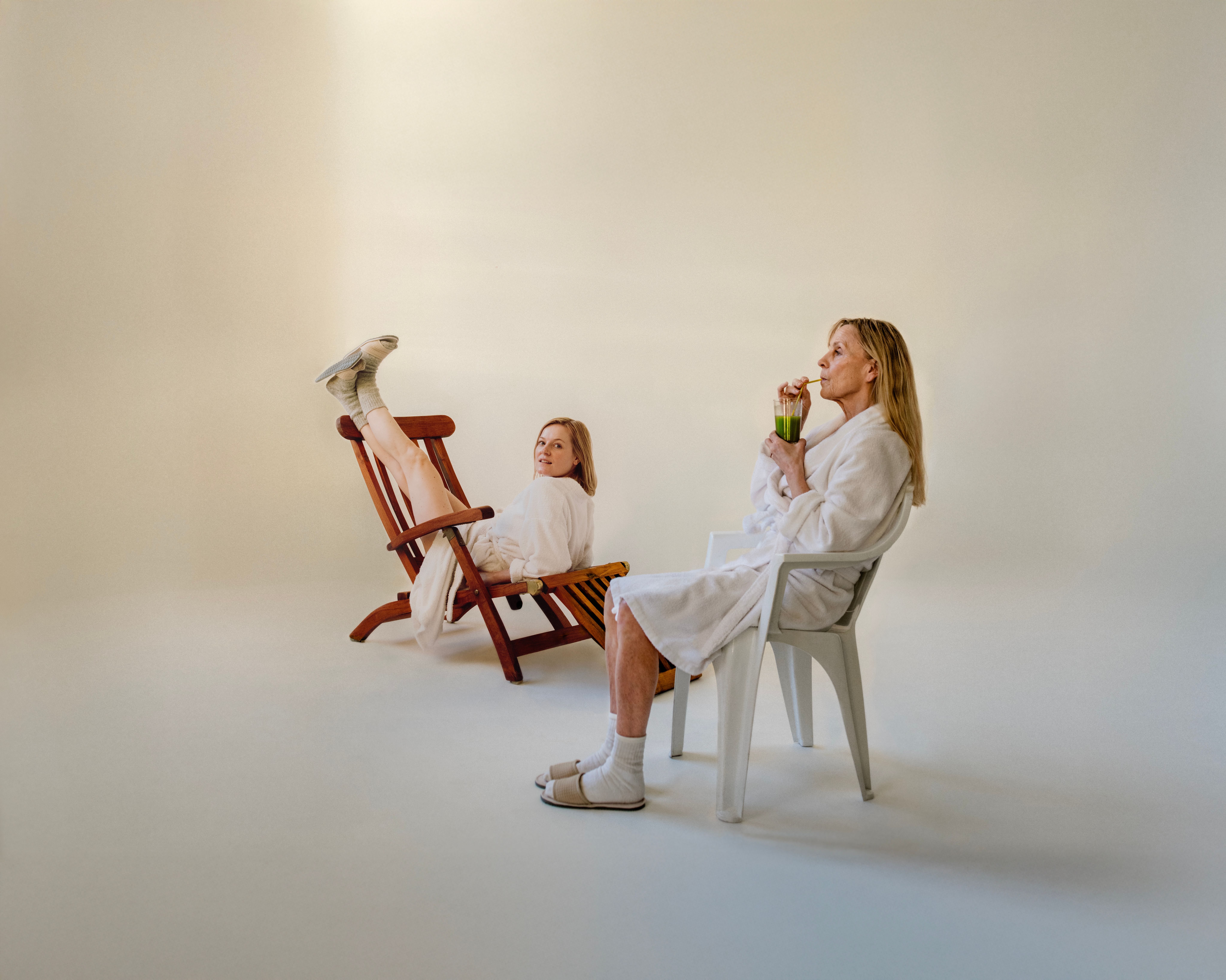 To kvinner i hvite morgenkåper. Den ene drikker en grønn jus mens hun sitter i en hvit plaststol. Den andre sitter feil vei med beina opp på en brun solseng mens hun ser mot kamera