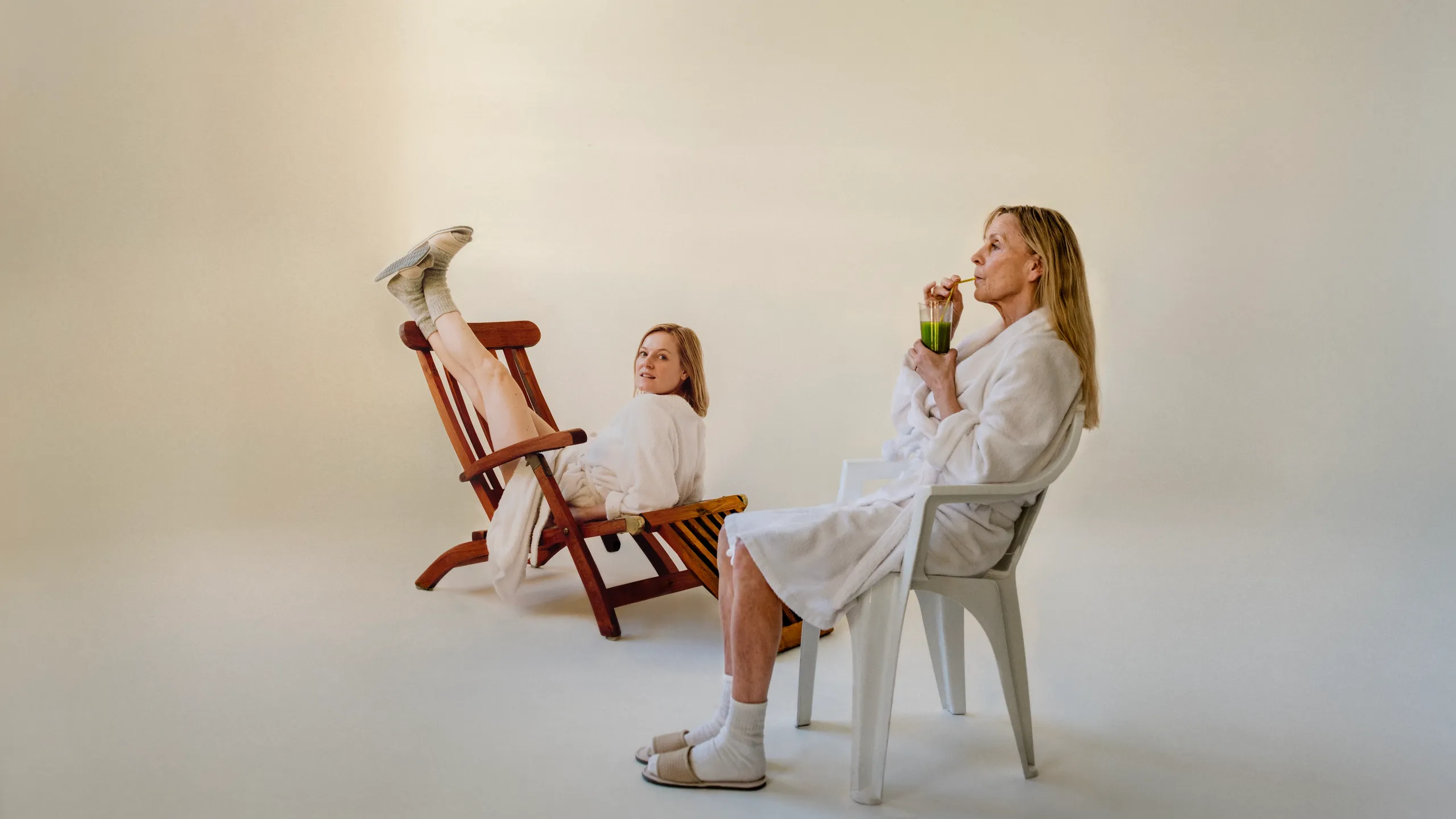 To kvinner i hvite morgenkåper. Den ene drikker en grønn jus mens hun sitter i en hvit plaststol. Den andre sitter feil vei med beina opp på en brun solseng mens hun ser mot kamera