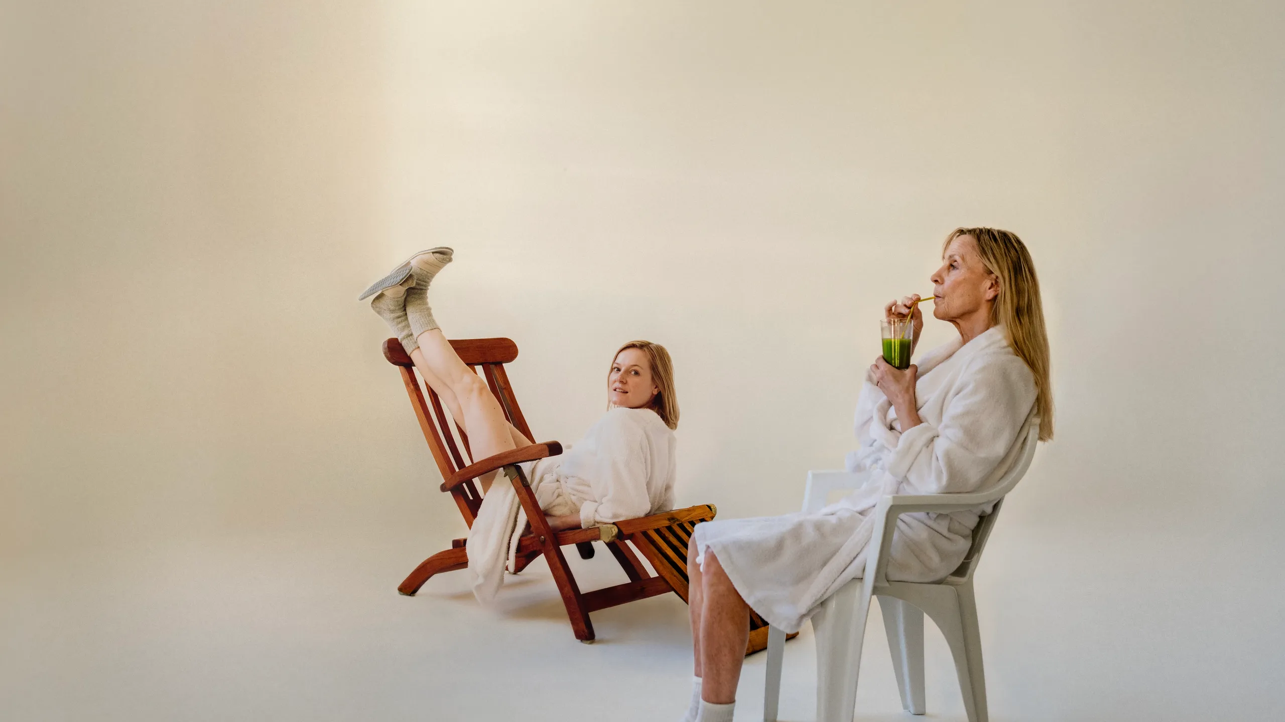 To kvinner i hvite morgenkåper. Den ene drikker en grønn jus mens hun sitter i en hvit plaststol. Den andre sitter feil vei med beina opp på en brun solseng mens hun ser mot kamera