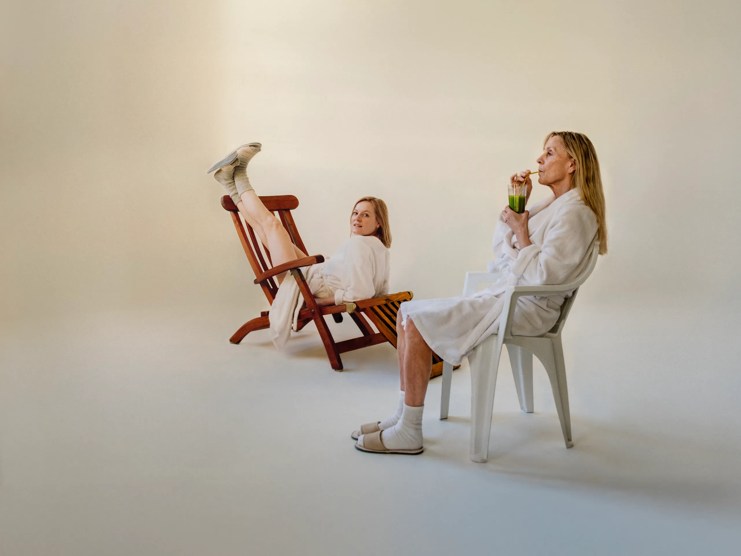 To kvinner i hvite morgenkåper. Den ene drikker en grønn jus mens hun sitter i en hvit plaststol. Den andre sitter feil vei med beina opp på en brun solseng mens hun ser mot kamera