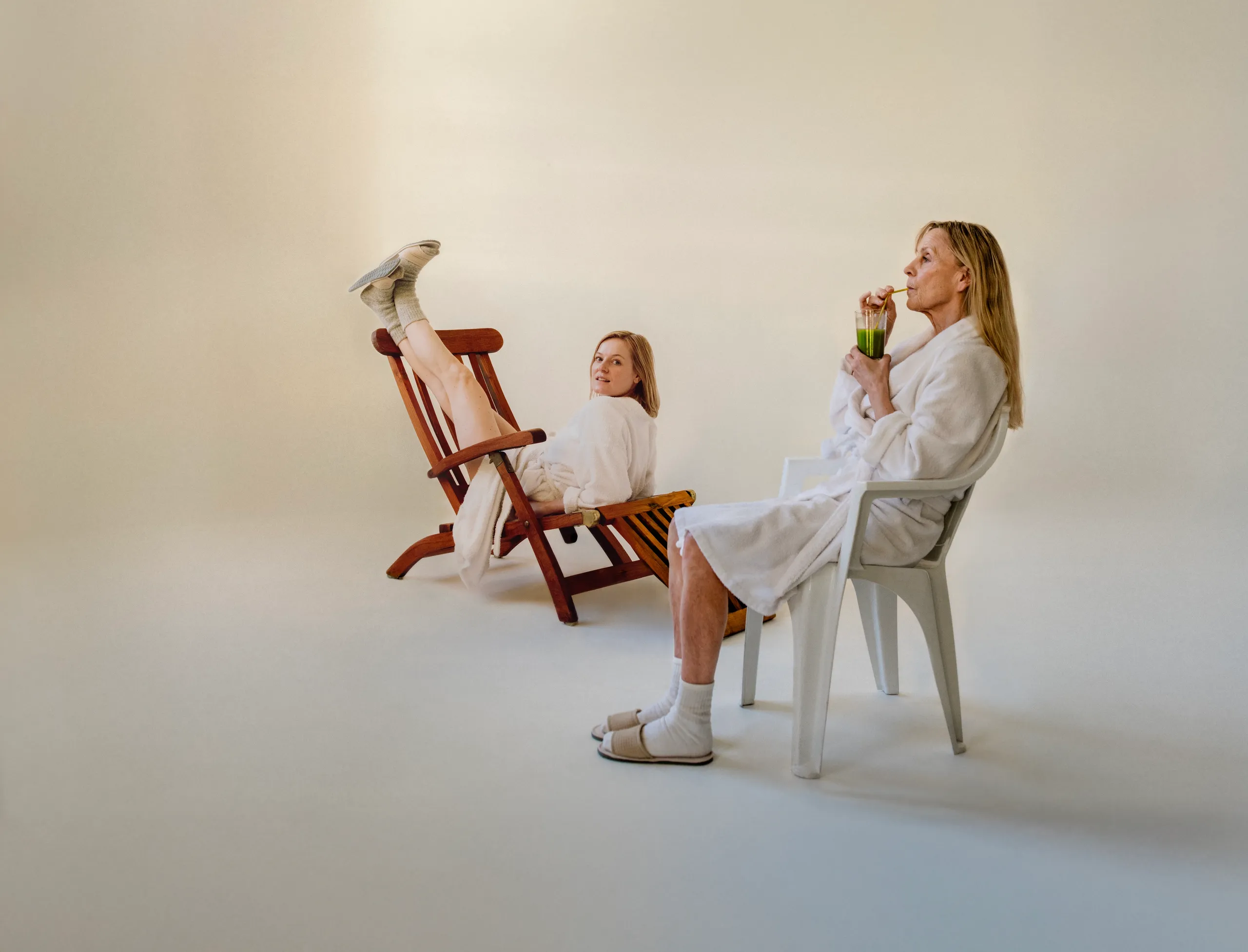 To kvinner i hvite morgenkåper. Den ene drikker en grønn jus mens hun sitter i en hvit plaststol. Den andre sitter feil vei med beina opp på en brun solseng mens hun ser mot kamera