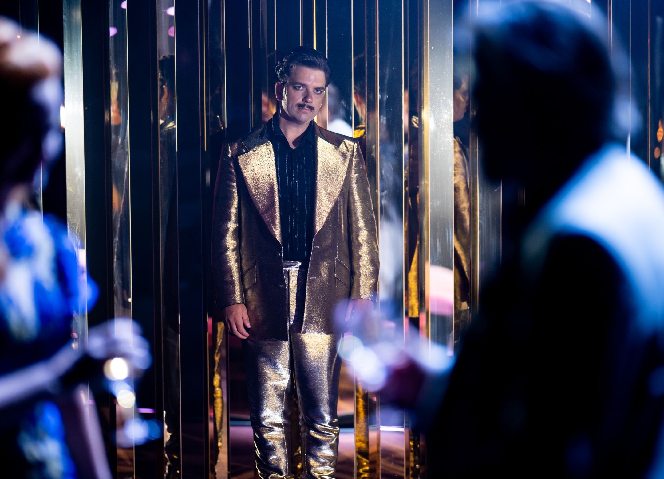 En mann i gullfarget dress og svart skjorte står foran speilvegger, med silhuetter av andre i forgrunnen.