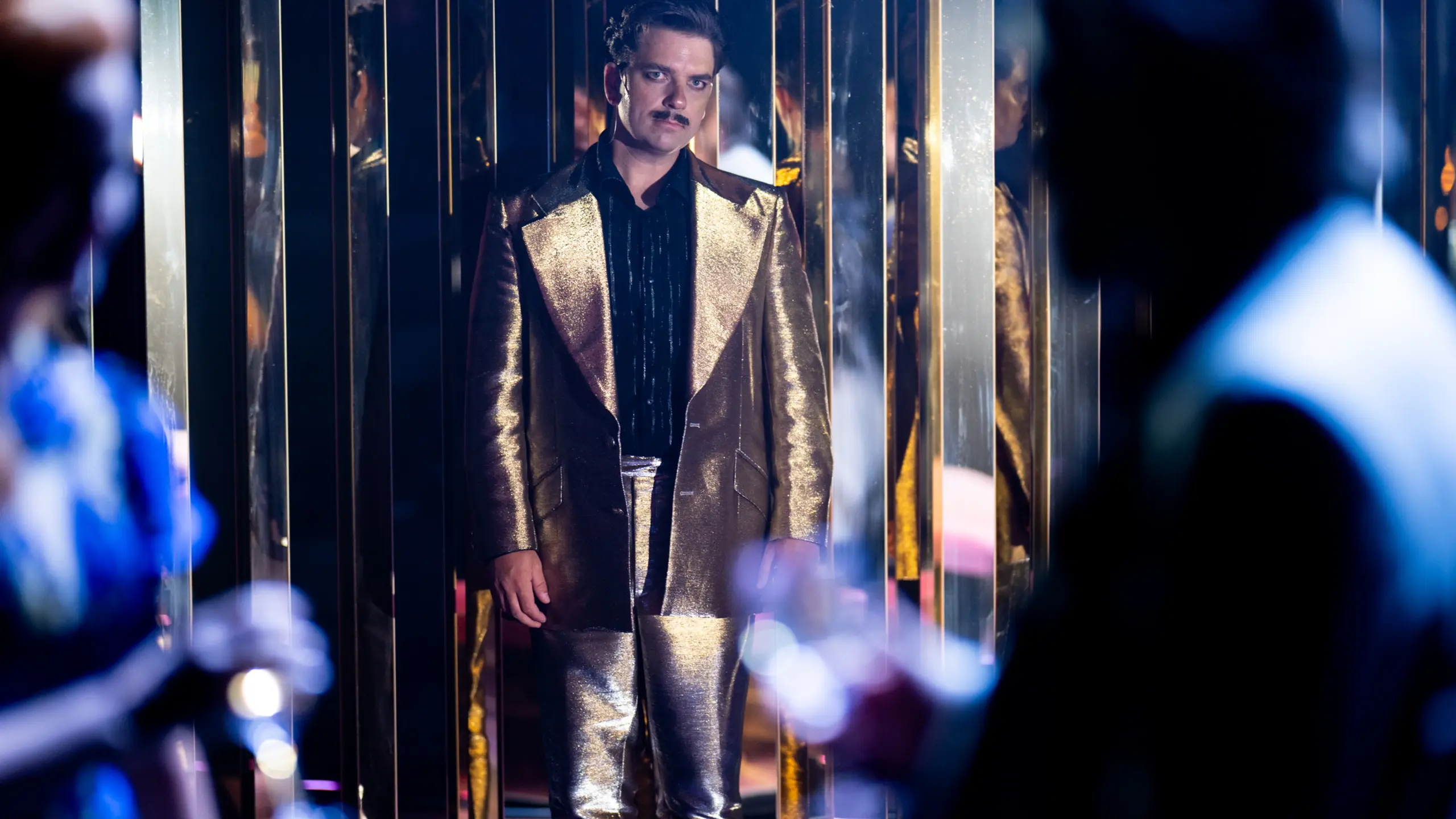 En mann i gullfarget dress og svart skjorte står foran speilvegger, med silhuetter av andre i forgrunnen.
