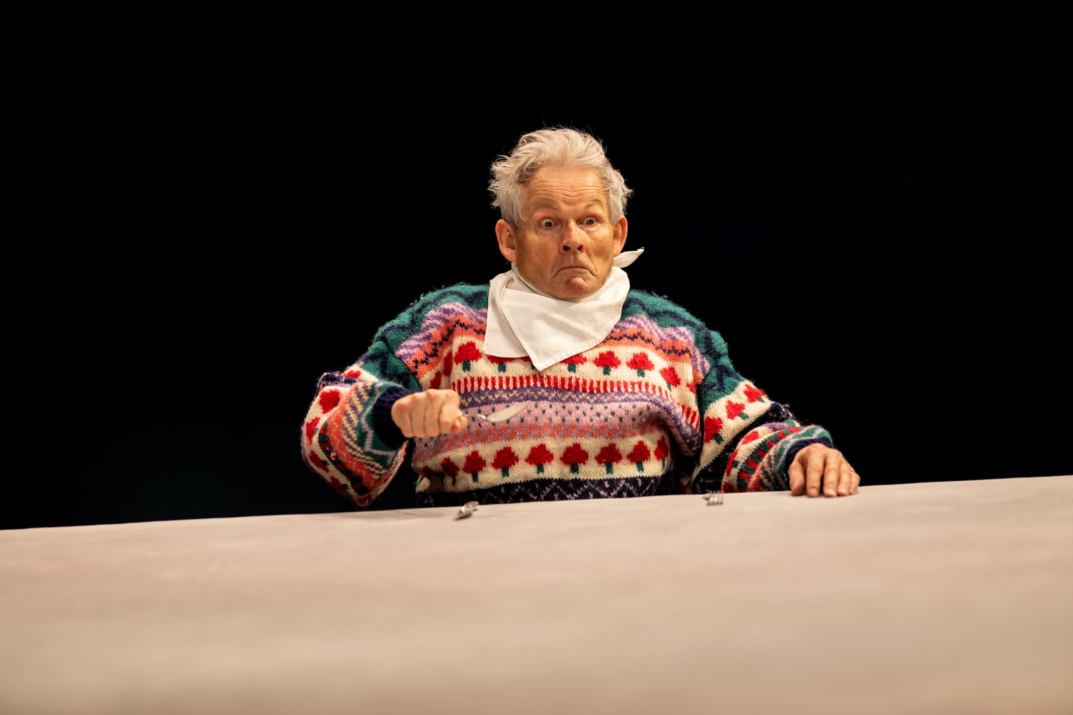 mann i fargerik strikkegenser med smekke sitter alene ved et bord og holder bestikk.