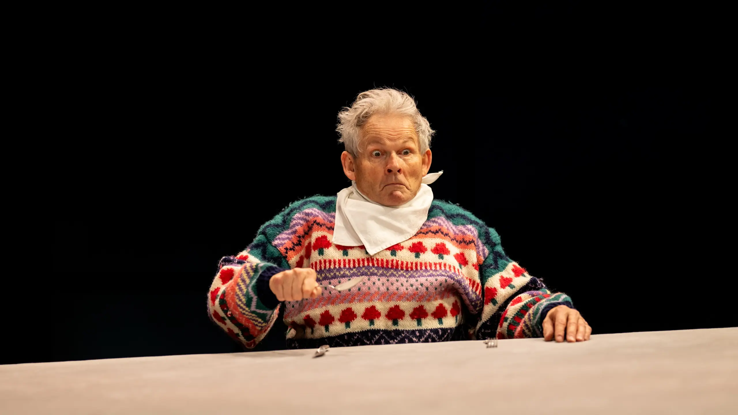 mann i fargerik strikkegenser med smekke sitter alene ved et bord og holder bestikk.