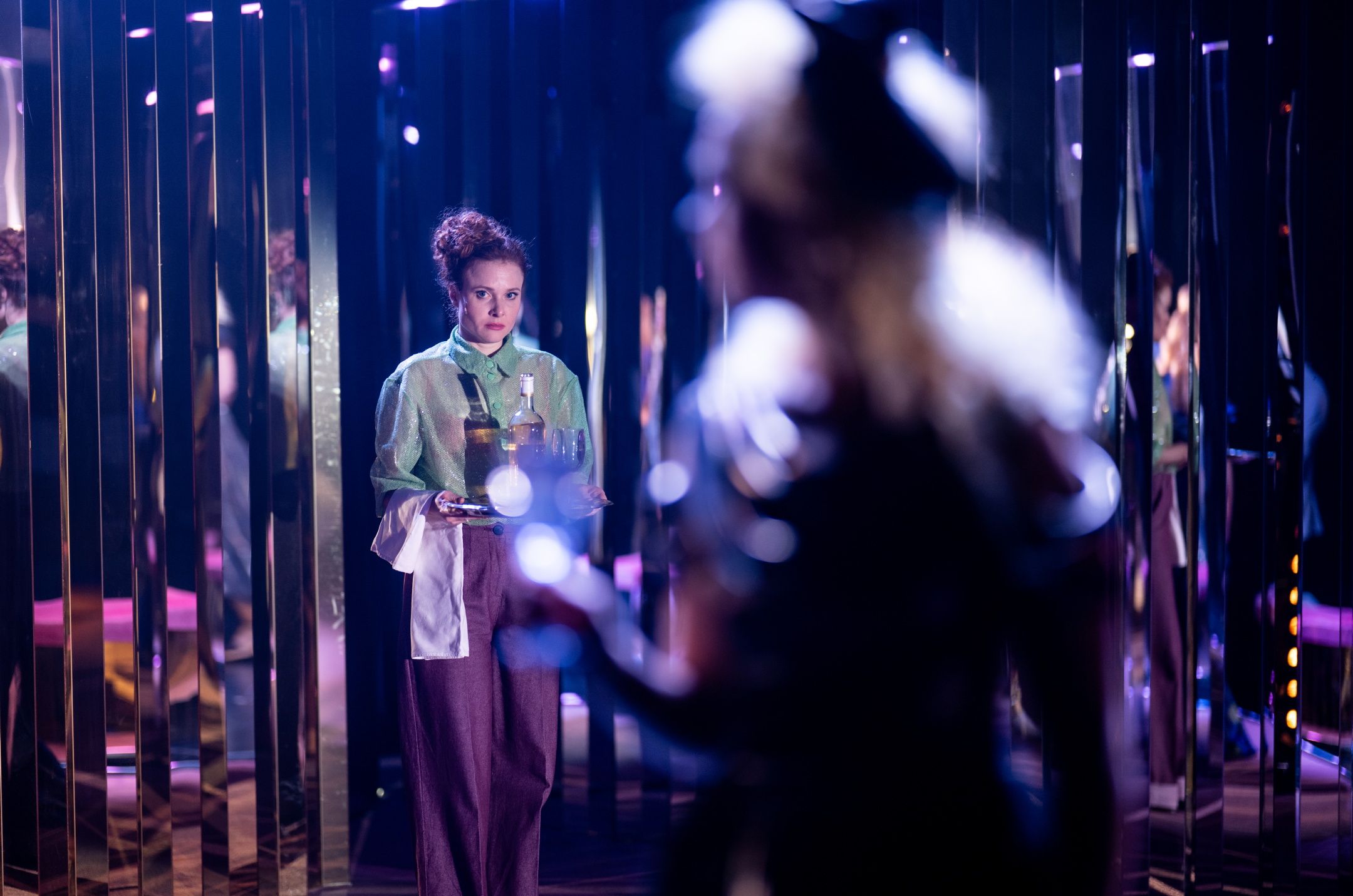 En kvinne i grønn skjorte og lilla bukser holder serveringsbrett med flasker og glass, omgitt av speilflater.