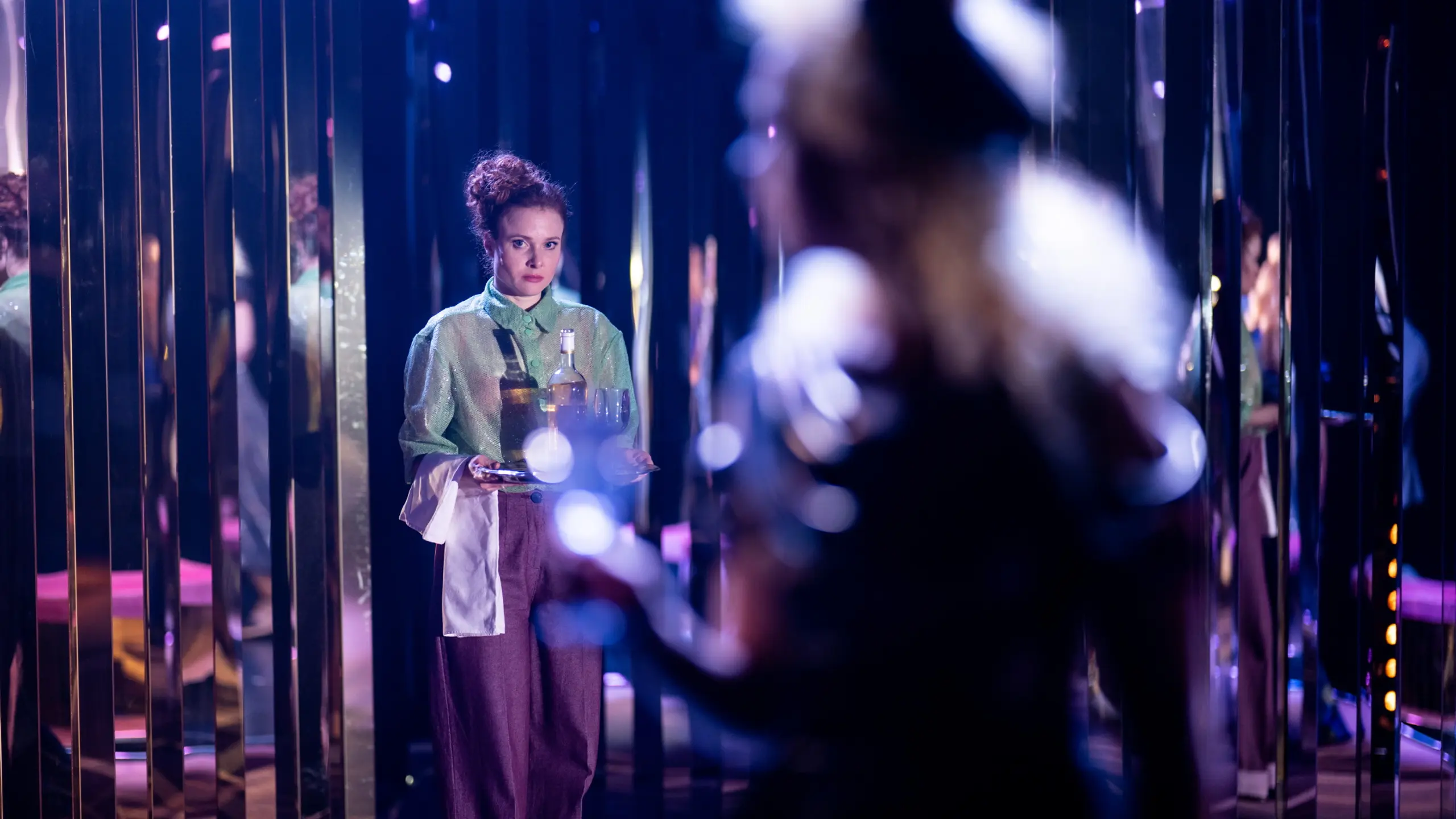 En kvinne i grønn skjorte og lilla bukser holder serveringsbrett med flasker og glass, omgitt av speilflater.