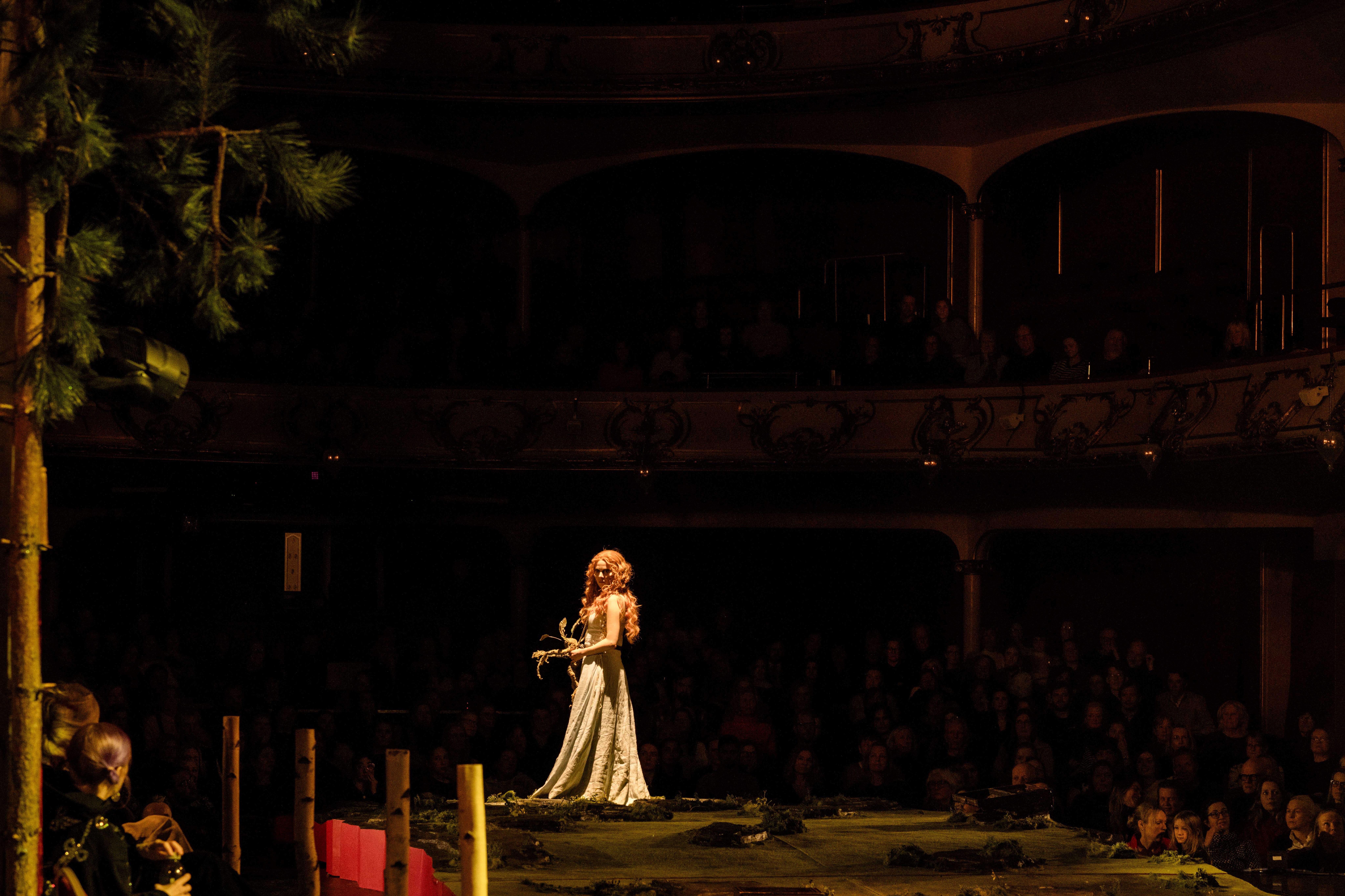 En kvinne i lang hvit kjole med langt rødt hår står alene på en scenografi med skogspreg, foran mørklagt sal.