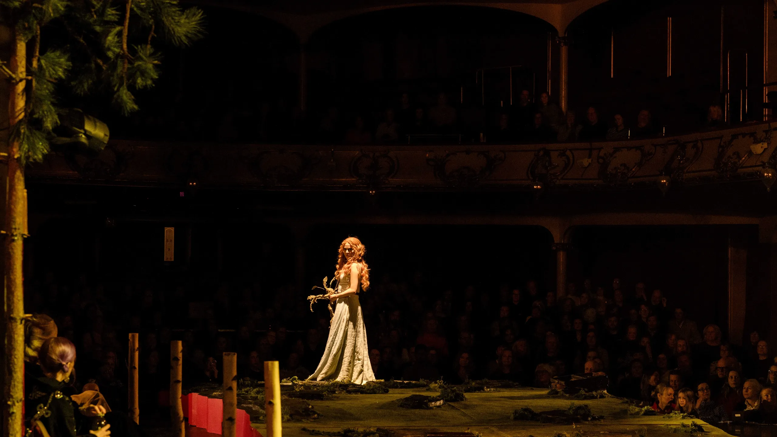 En kvinne i lang hvit kjole med langt rødt hår står alene på en scenografi med skogspreg, foran mørklagt sal.