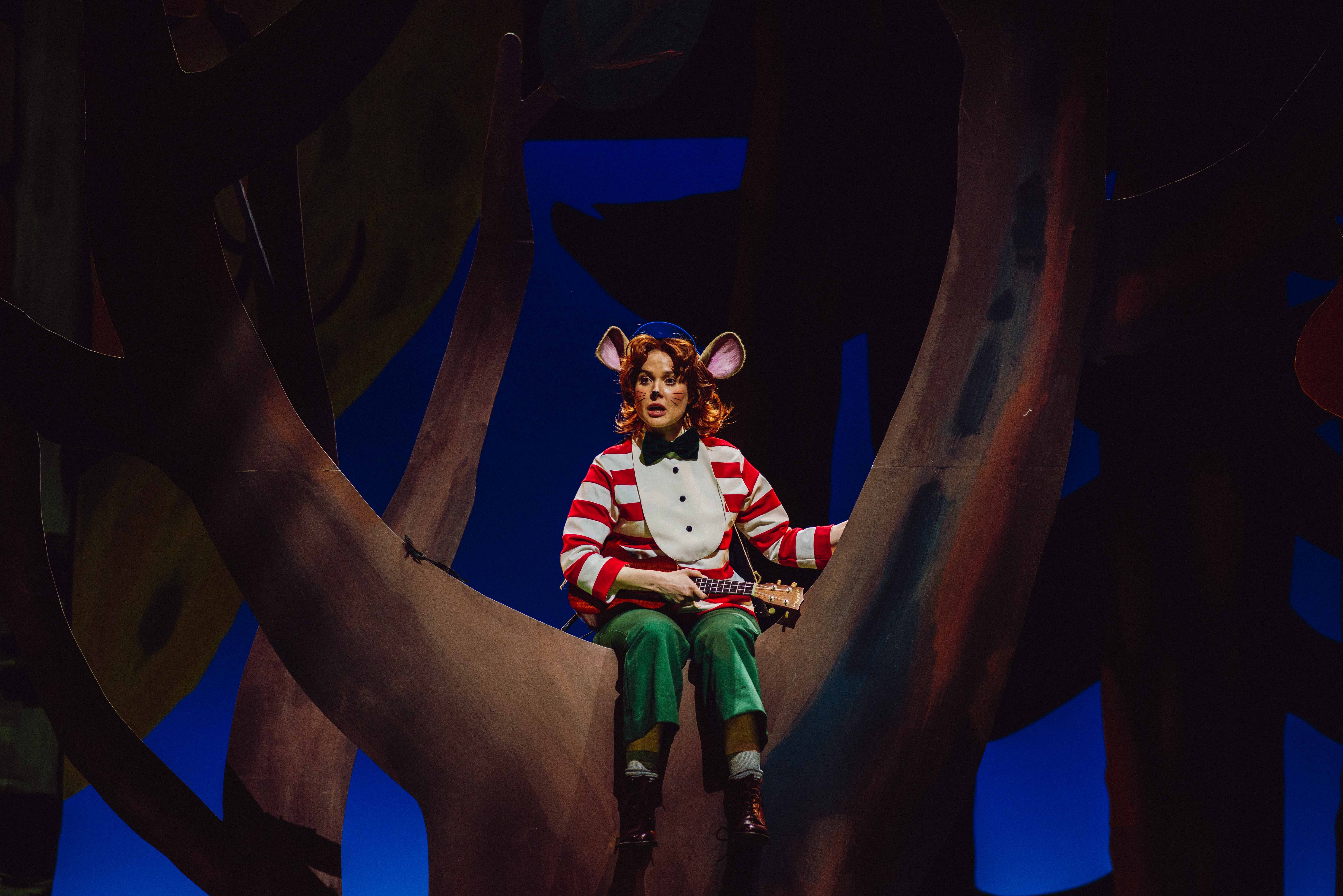 Mus i røde striper og grønn bukse sitter i et tre og spiller ukulele.