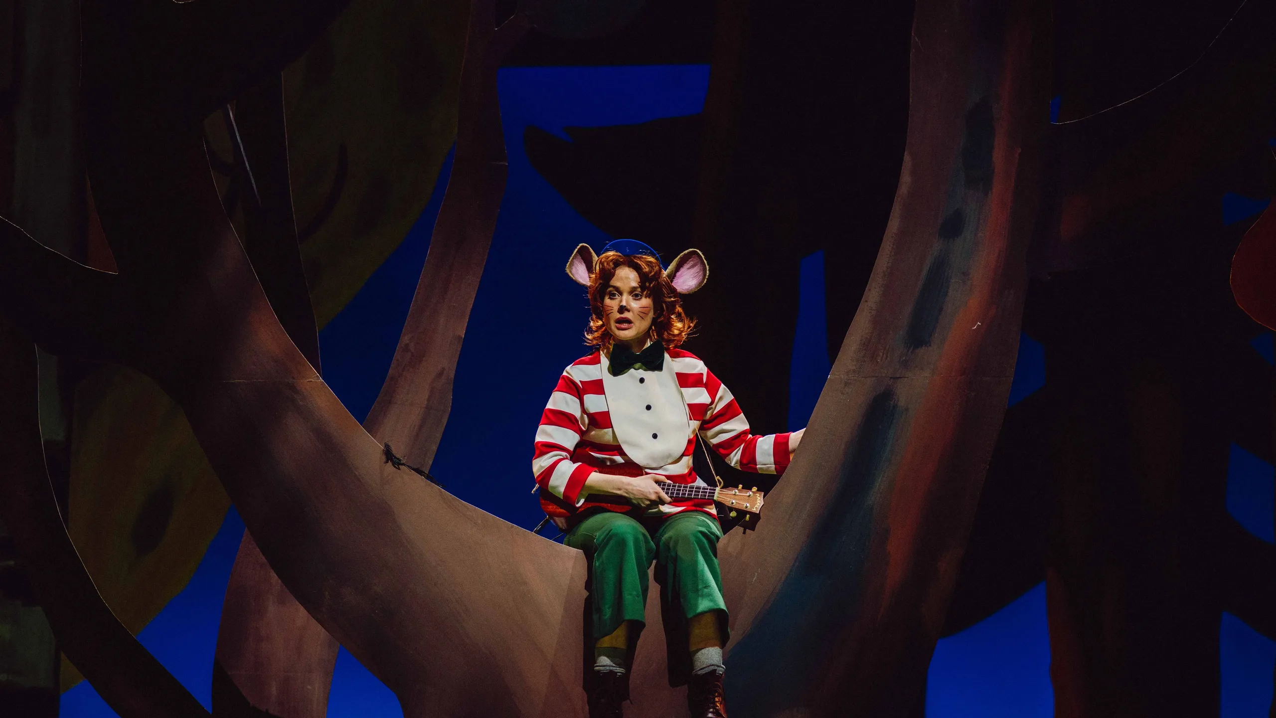 Mus i røde striper og grønn bukse sitter i et tre og spiller ukulele.