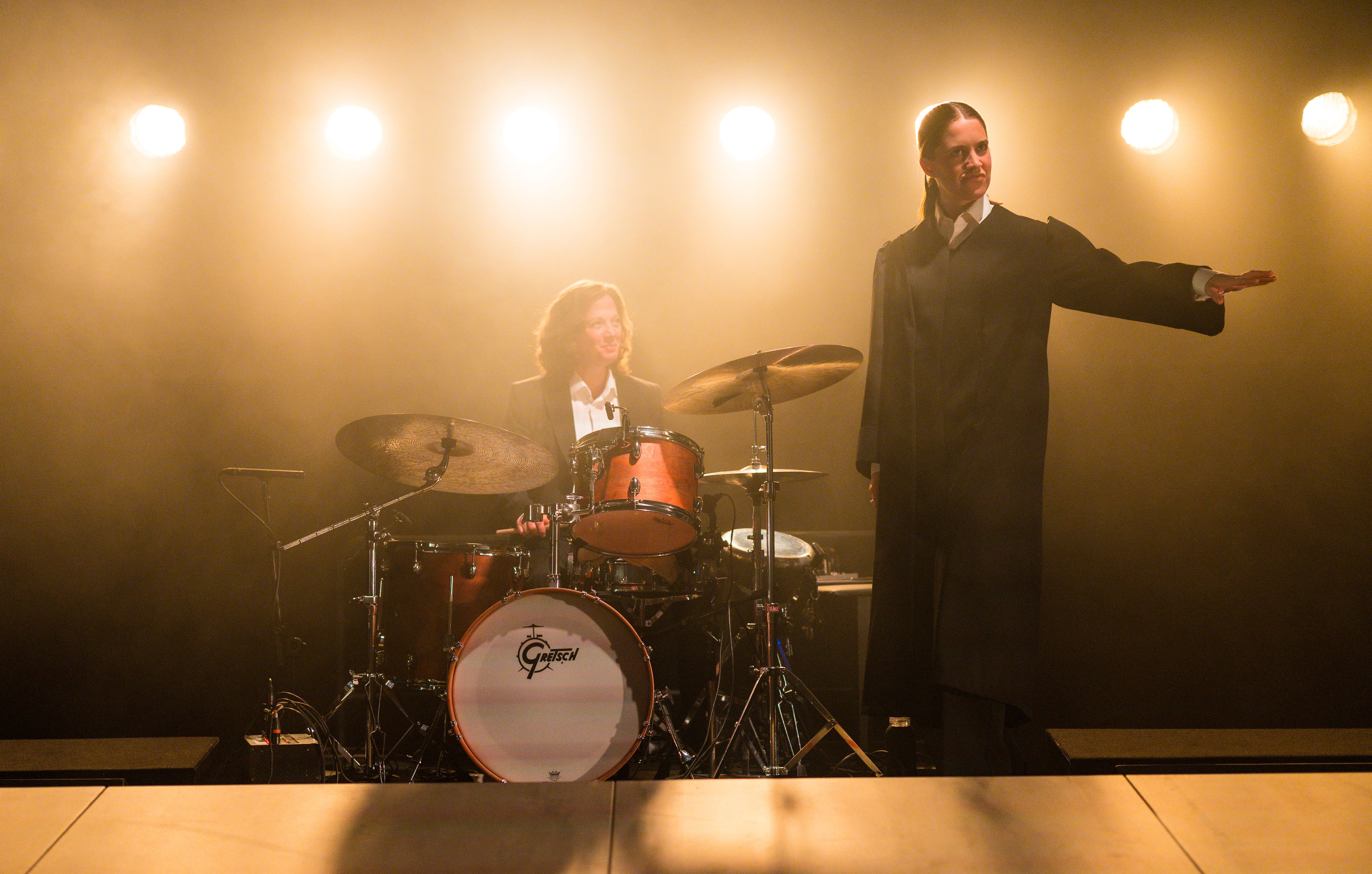 En kvinne i svart kåpe står ved siden av en trommeslager på scenen.