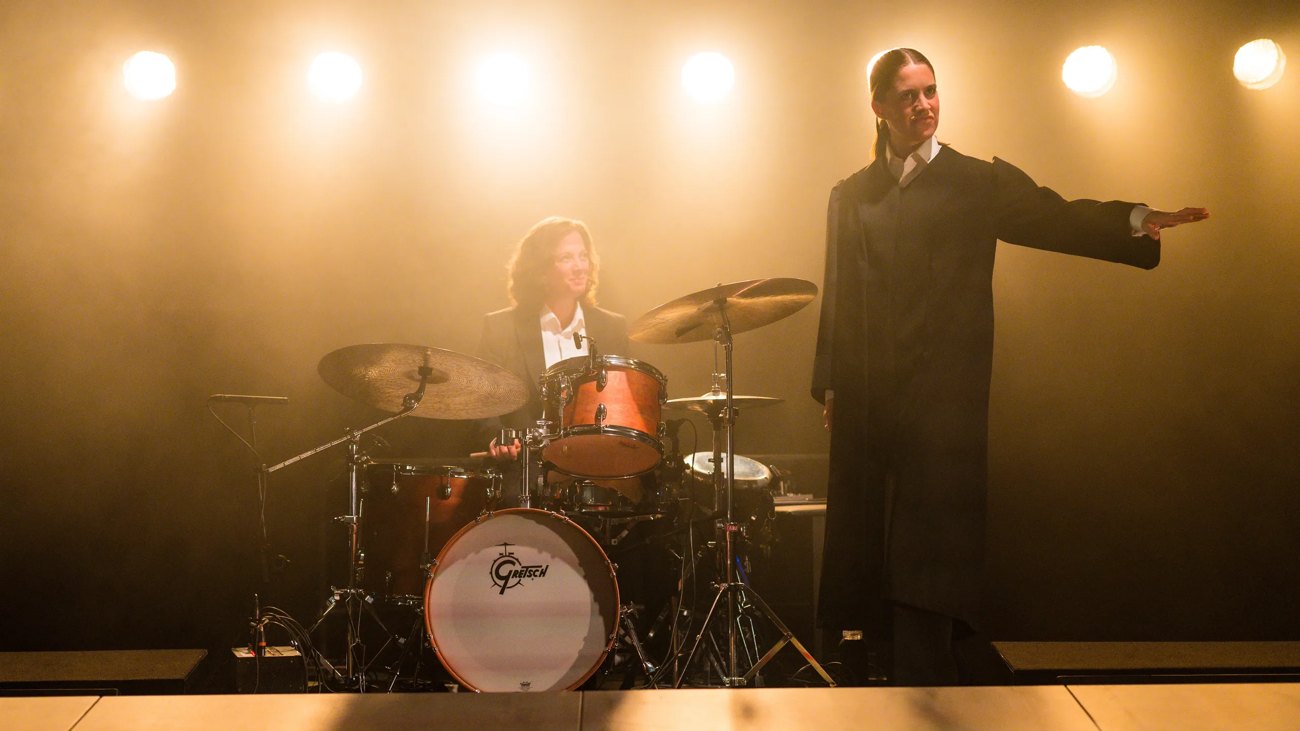 En kvinne i svart kåpe står ved siden av en trommeslager på scenen.