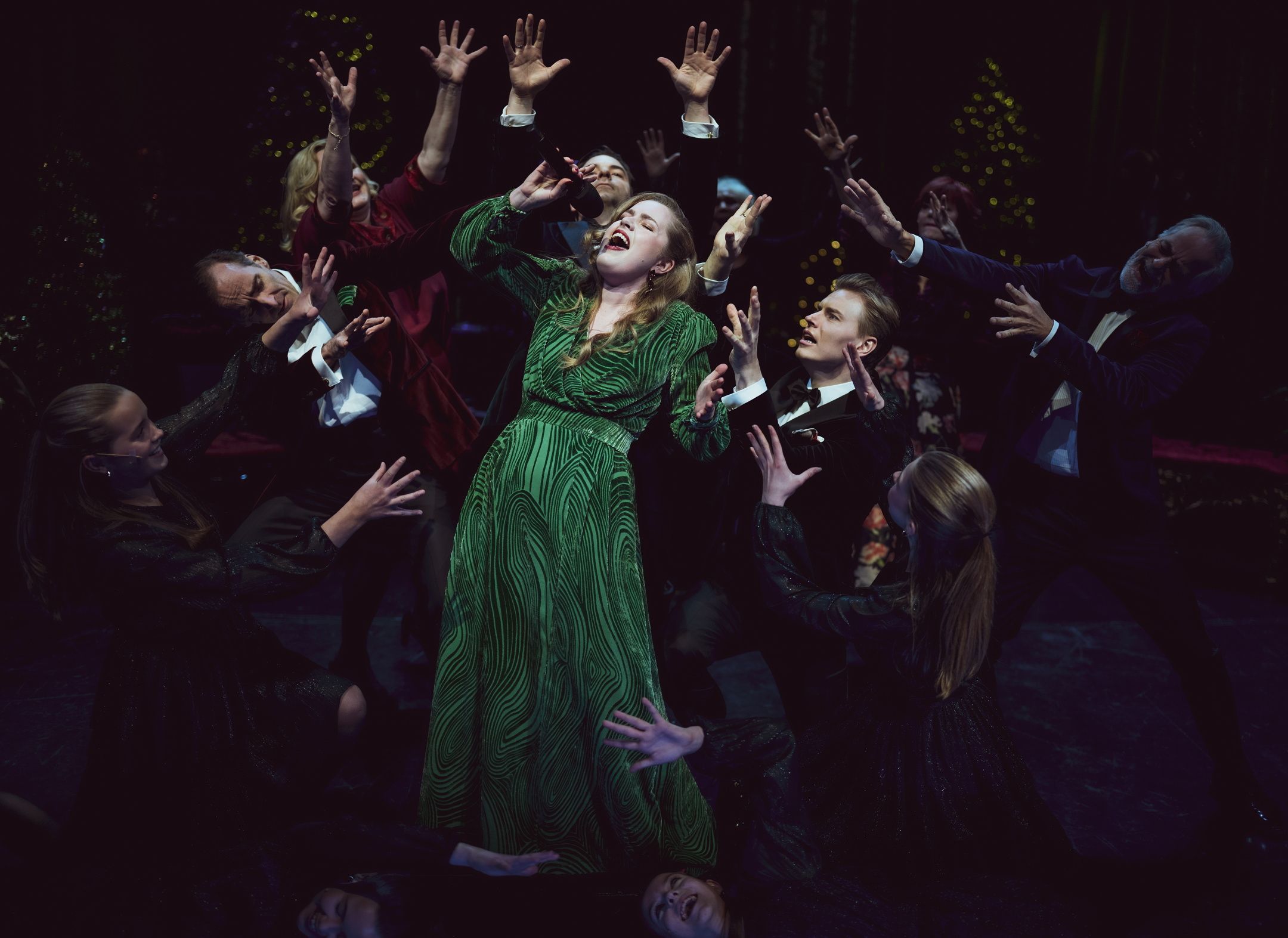 Kvinne synger, mens en gruppe artister står rundt og strekker armene mot henne. Alle er festkledd