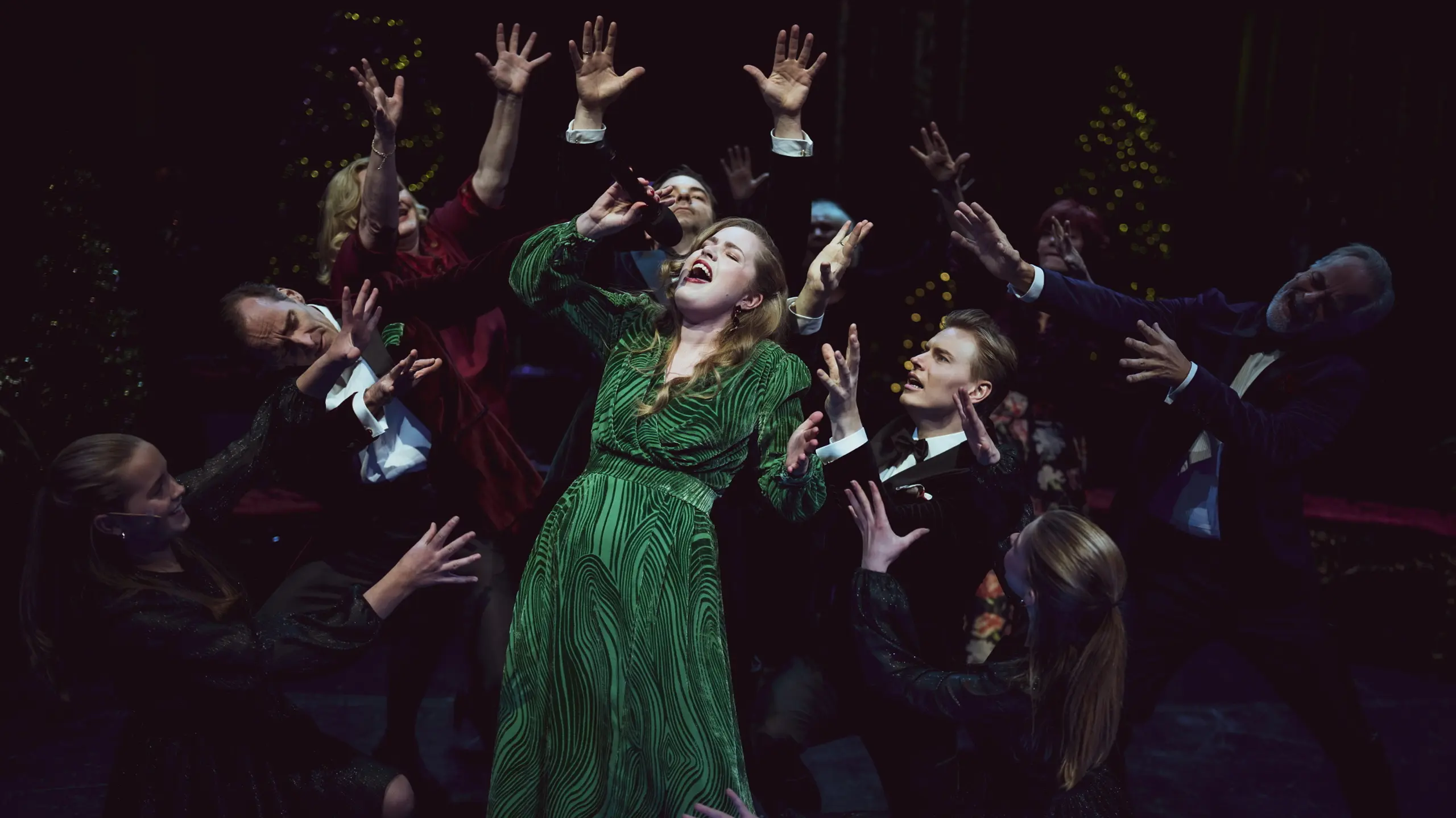 Kvinne synger, mens en gruppe artister står rundt og strekker armene mot henne. Alle er festkledd