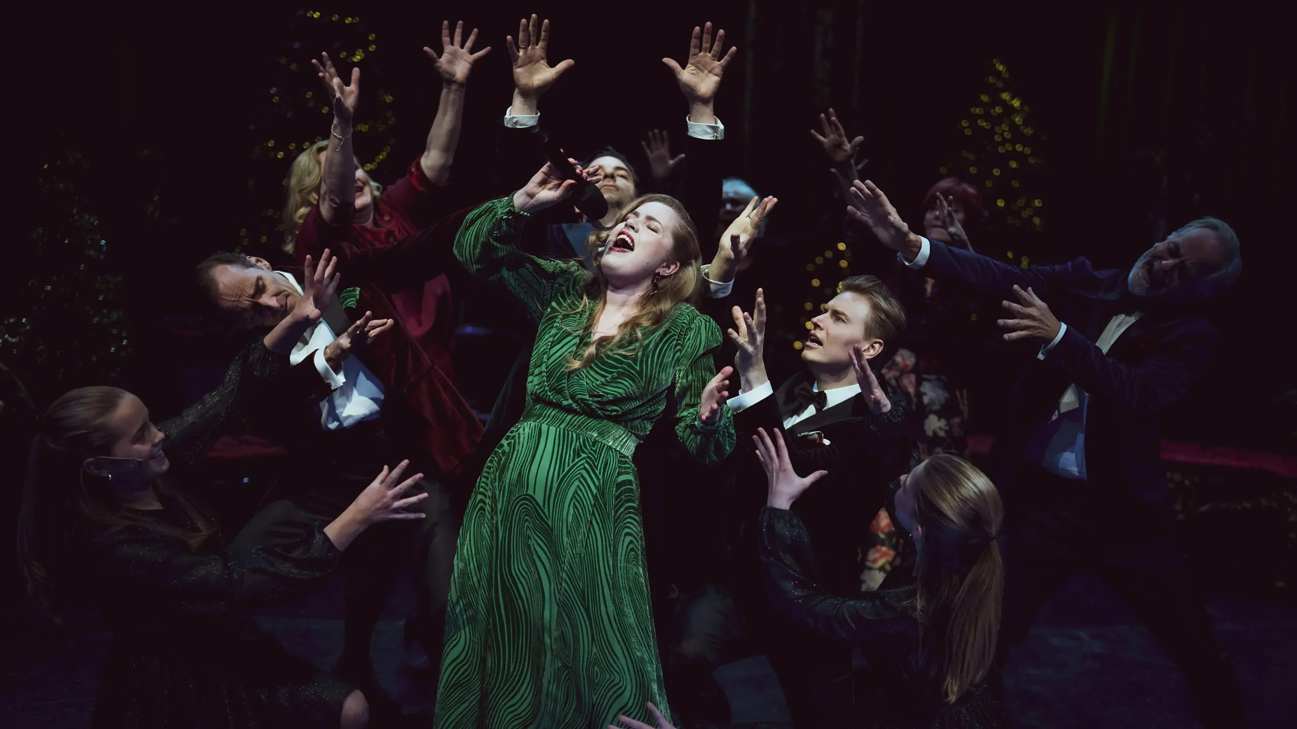 Kvinne synger, mens en gruppe artister står rundt og strekker armene mot henne. Alle er festkledd