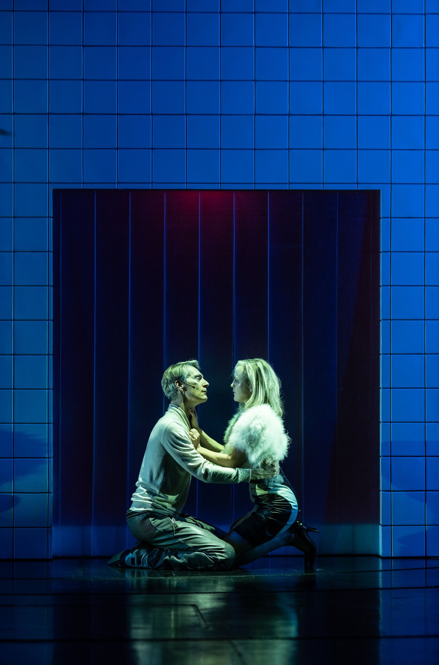 Par kneler foran hverandre i scene omgitt av blå fliser; kvinnen har hvit pelsjakke, mannen lys genser.