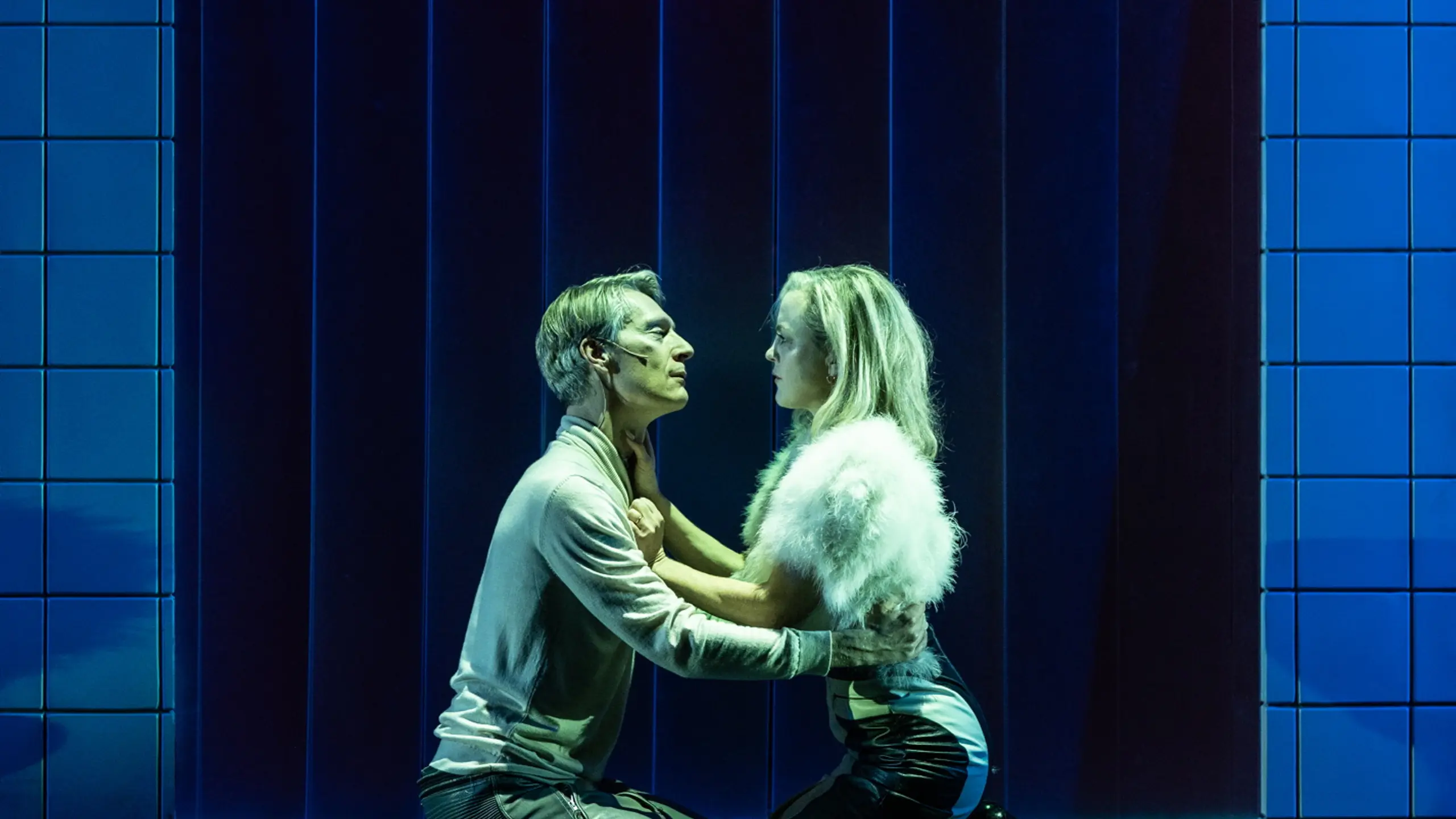 Par kneler foran hverandre i scene omgitt av blå fliser; kvinnen har hvit pelsjakke, mannen lys genser.