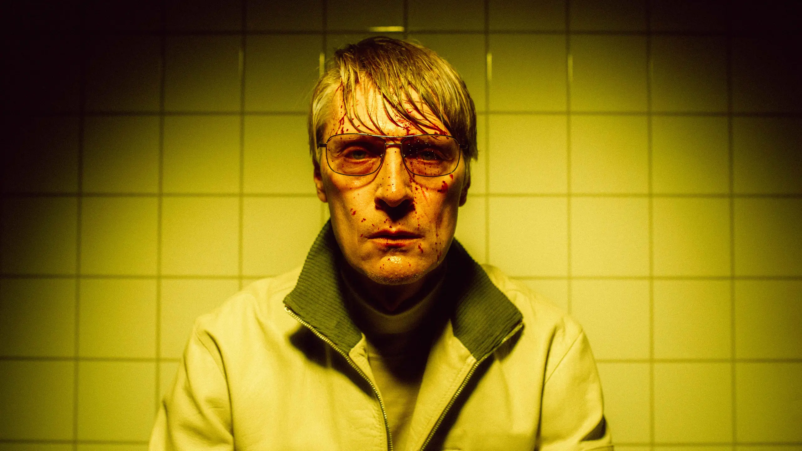 Svett mann med solbriller og blodsprut i fjeset i gult lys. Hvite fliser på veggen bak