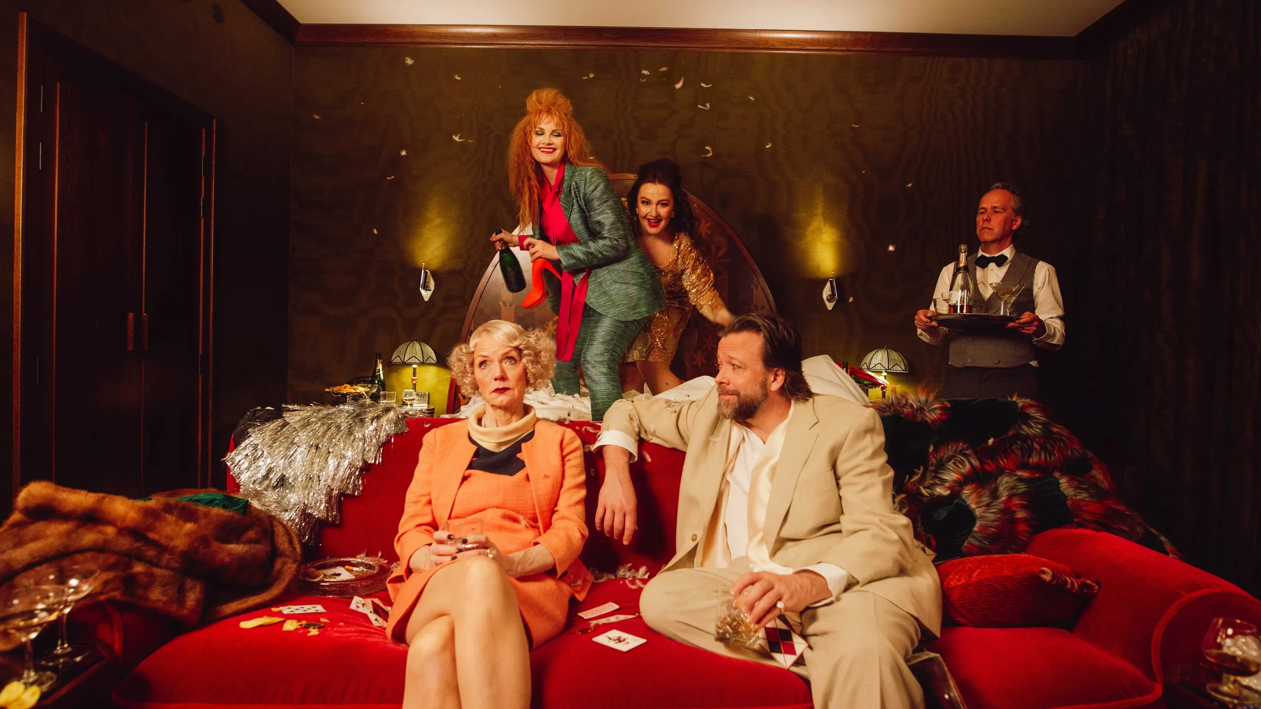 Fire skuespillere som herjer på et hotellrom. To kvinner hopper i senga, mens en kvinne og en mann sitter i sofaen