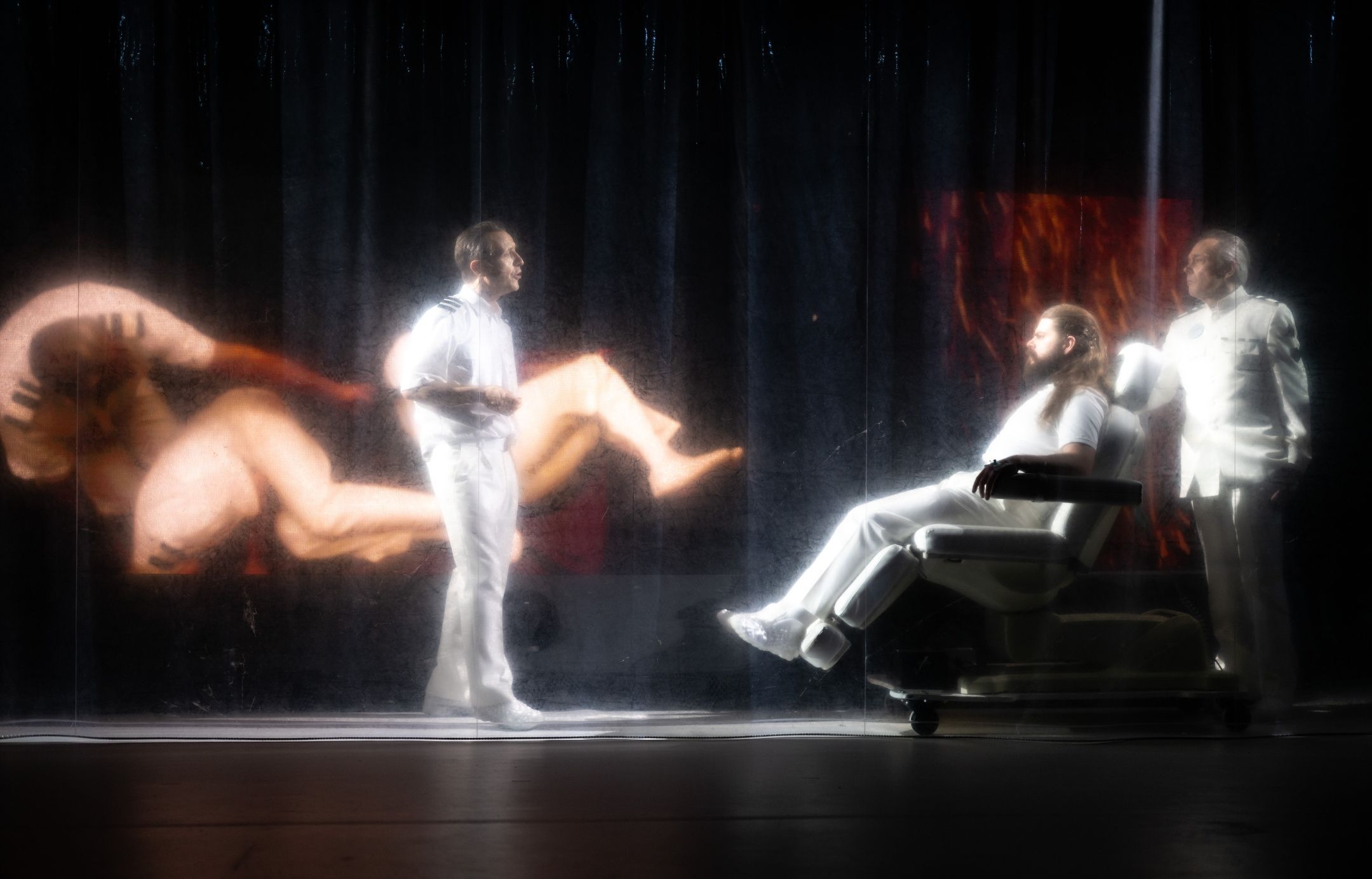 To personer i hvite uniformer står ved siden av en person i en stol, projisert naken figur vises på bakveggen