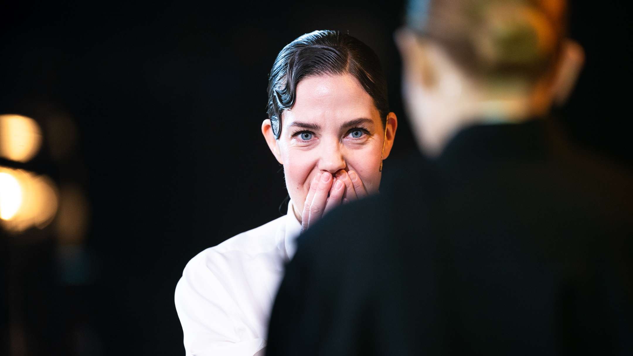 En kvinne i hvit skjorte dekker munnen med hendene i et uttrykk av overraskelse eller glede. Hun ser mot en annen person som er vendt bort fra kamera. Lyset bak henne gir scenen en varm og intim følelse.