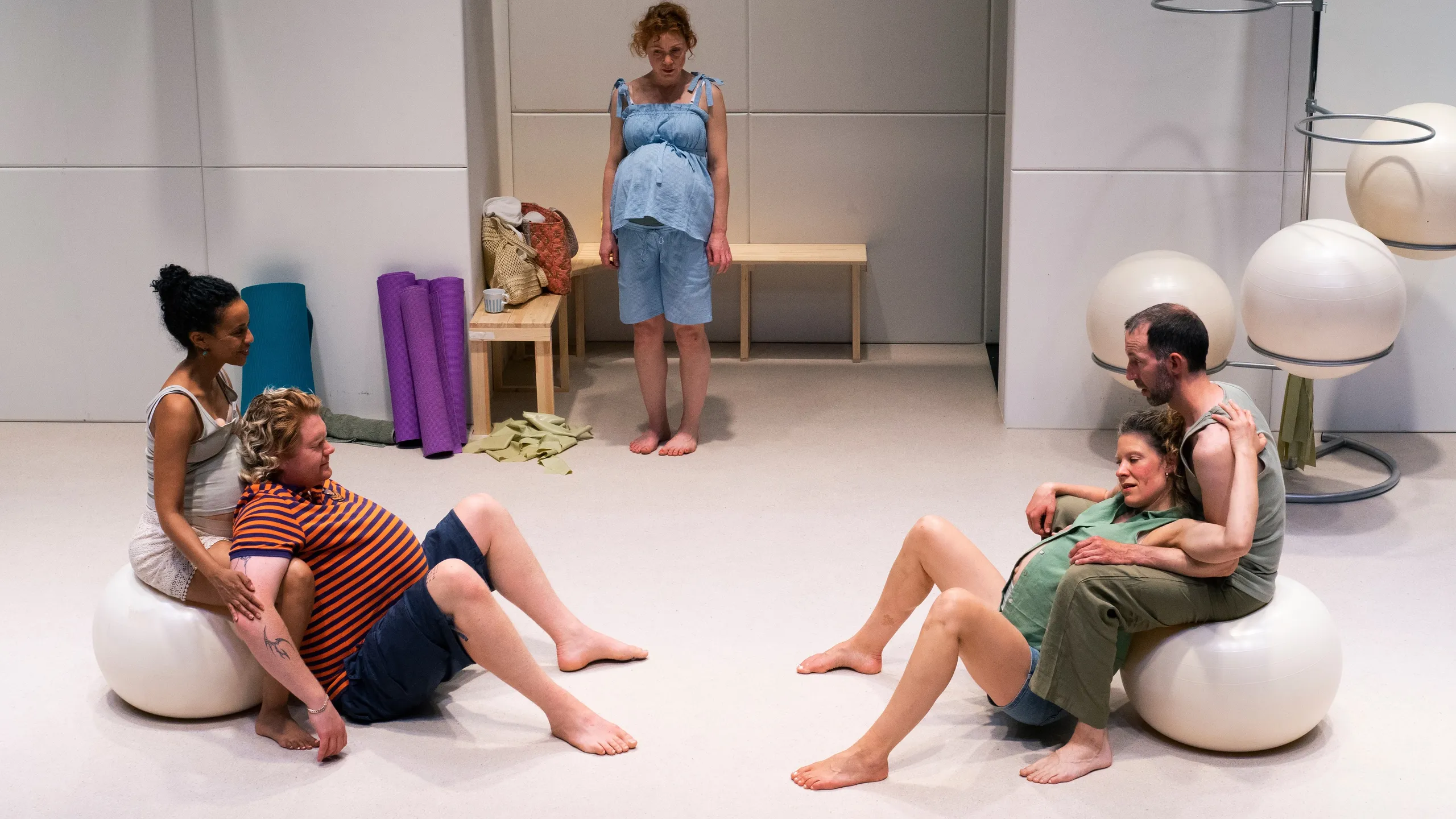 Fem personer i treningsrom, togravide par sitter på pilatesballer eller gulvet, én kvinne står i bakgrunnen.