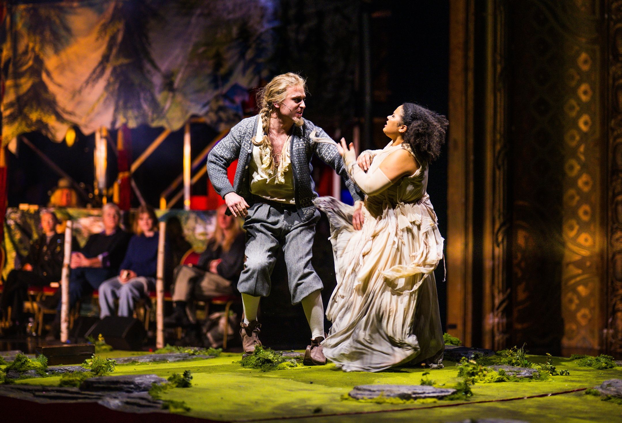 En mann i tradisjonell vestlandsdrakt og en kvinne i hvit kjole gestikulerer dramatisk mot hverandre foran et publikum.