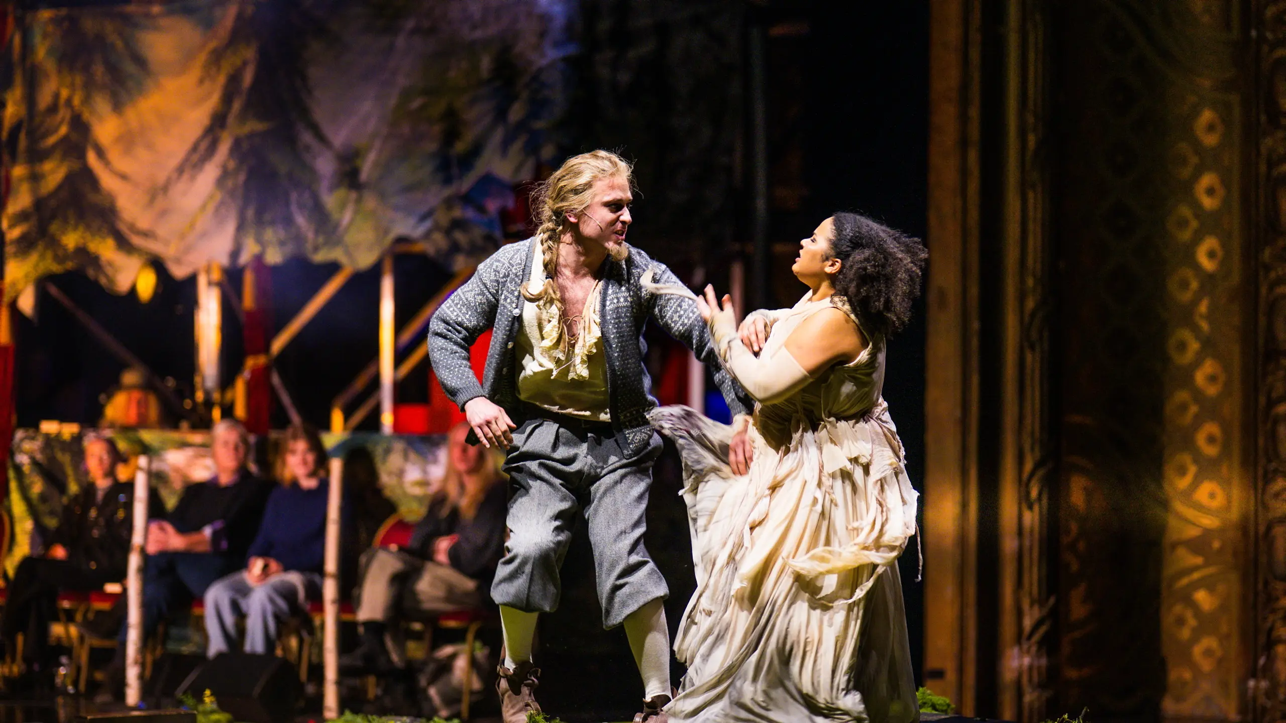 En mann i tradisjonell vestlandsdrakt og en kvinne i hvit kjole gestikulerer dramatisk mot hverandre foran et publikum.