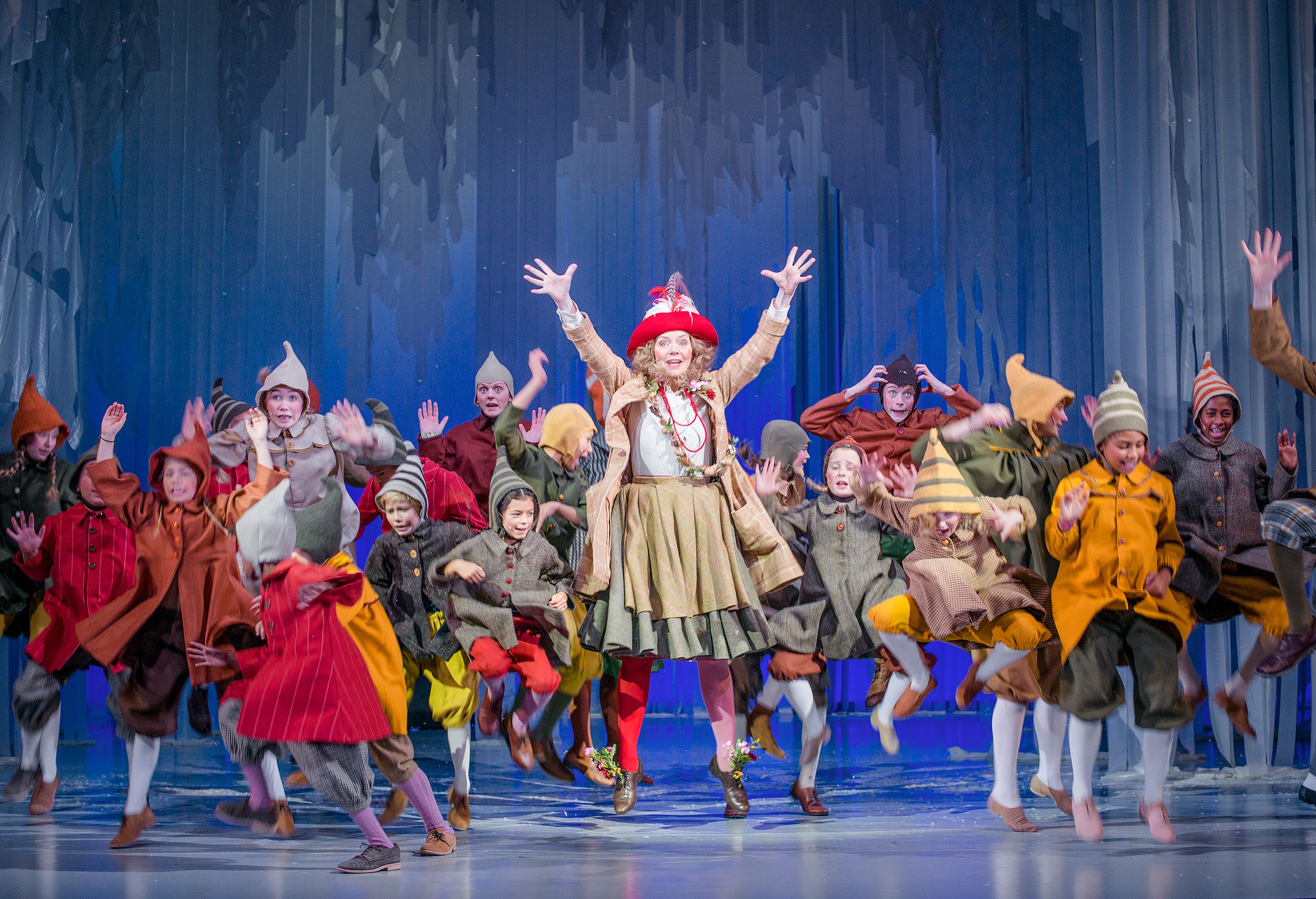 Masse barn og en kvinne i fargerike nisseaktige kostymer hopper og danser rundt på en scene med blå kulisser