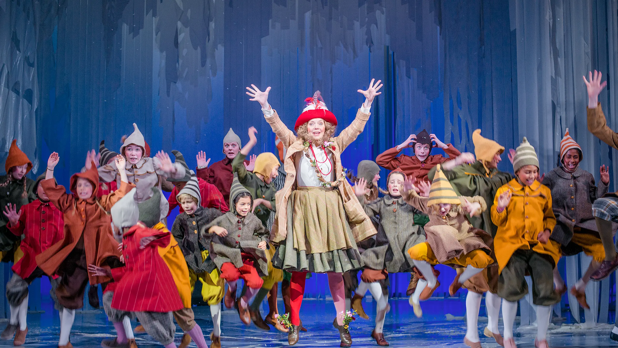 Masse barn og en kvinne i fargerike nisseaktige kostymer hopper og danser rundt på en scene med blå kulisser