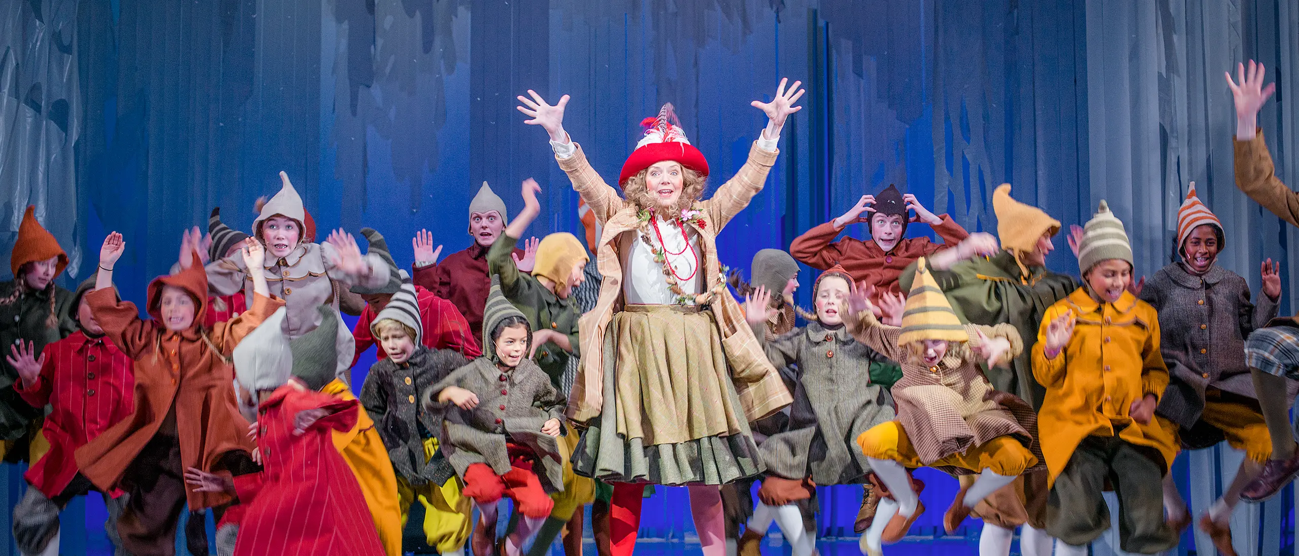 Masse barn og en kvinne i fargerike nisseaktige kostymer hopper og danser rundt på en scene med blå kulisser