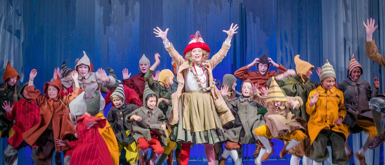 Masse barn og en kvinne i fargerike nisseaktige kostymer hopper og danser rundt på en scene med blå kulisser