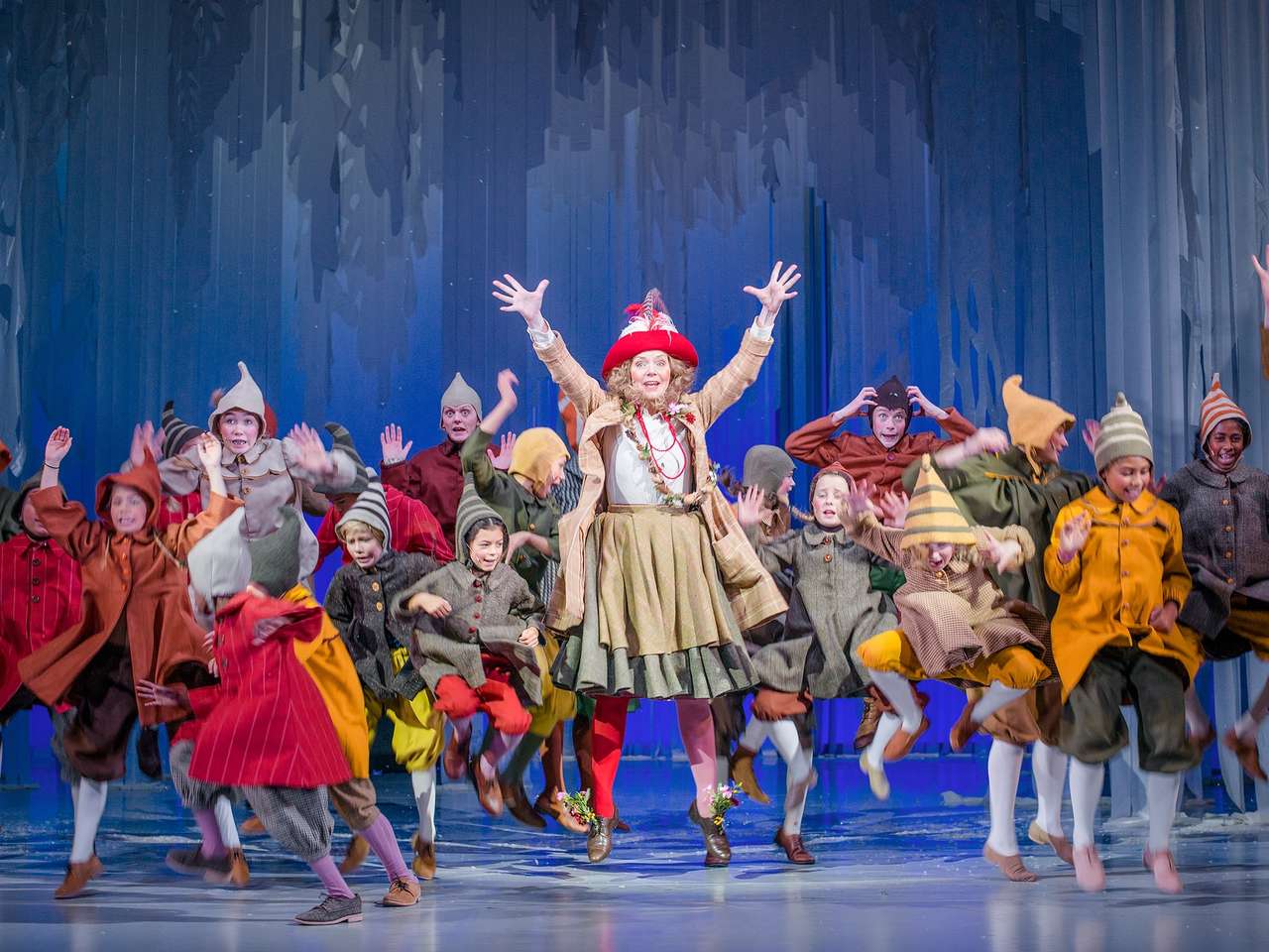 Masse barn og en kvinne i fargerike nisseaktige kostymer hopper og danser rundt på en scene med blå kulisser