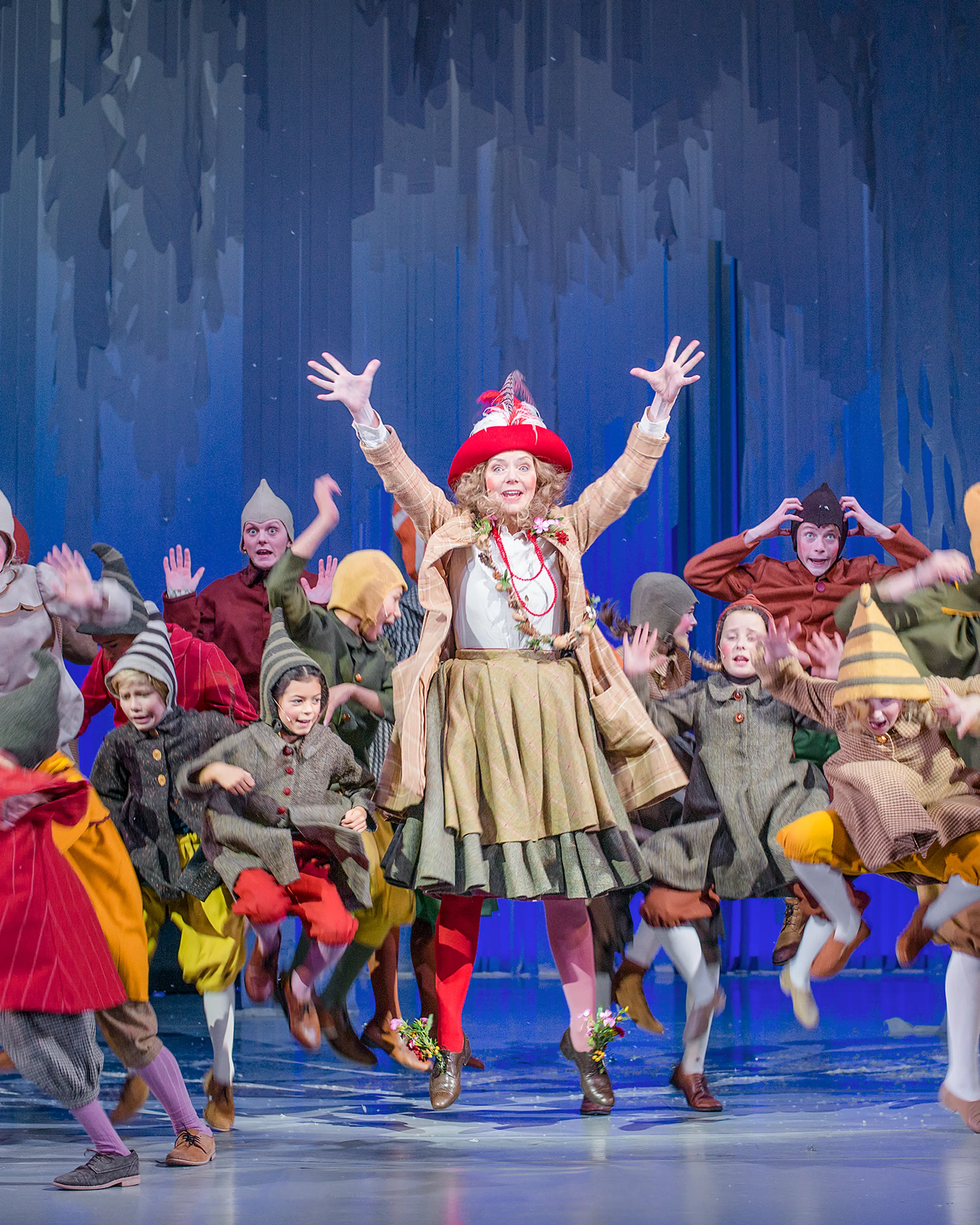 Masse barn og en kvinne i fargerike nisseaktige kostymer hopper og danser rundt på en scene med blå kulisser